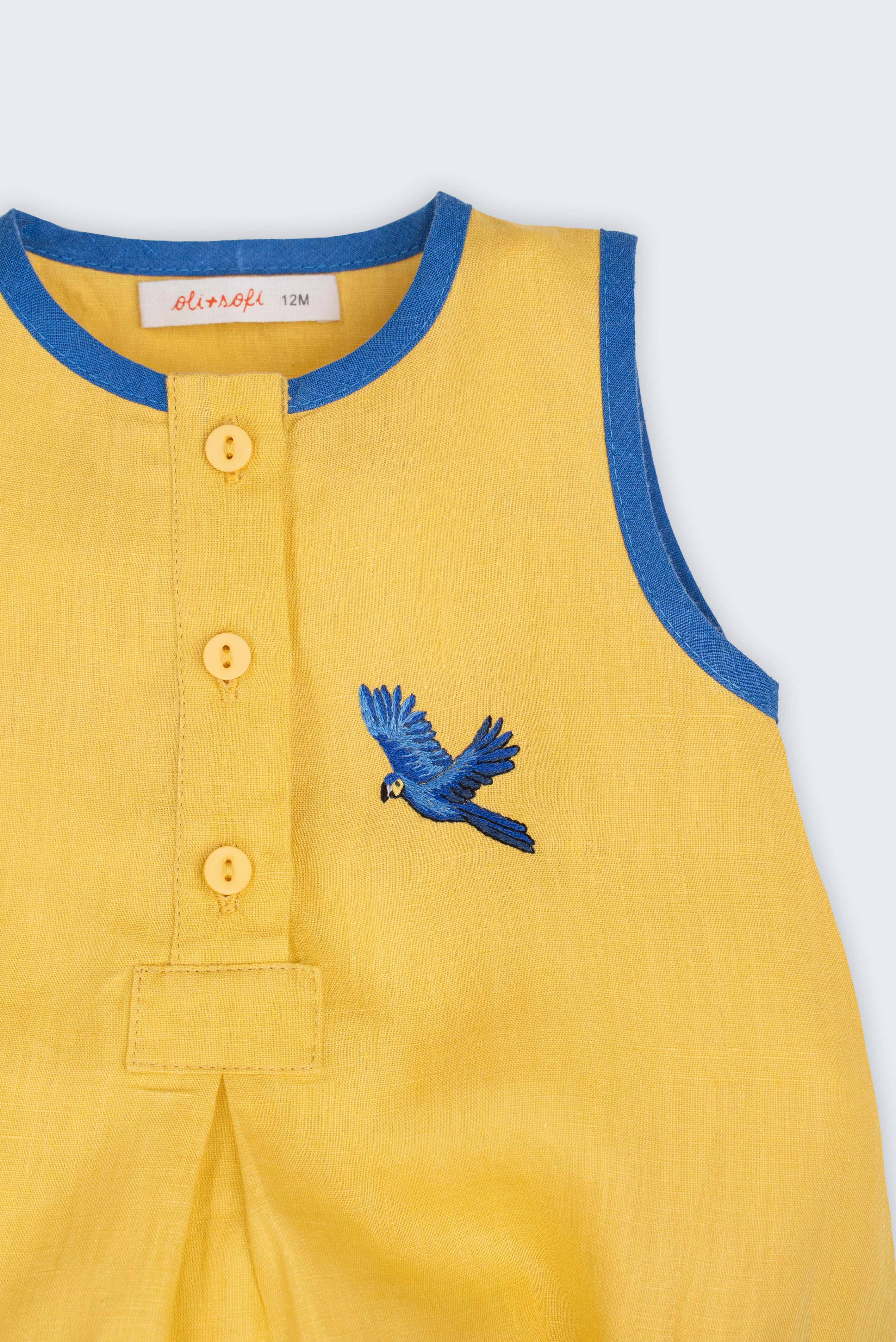 Macaquinho bebê em linho amarelo com acabamento azul e arara azul bordada à mão no peito, visto em detalhe