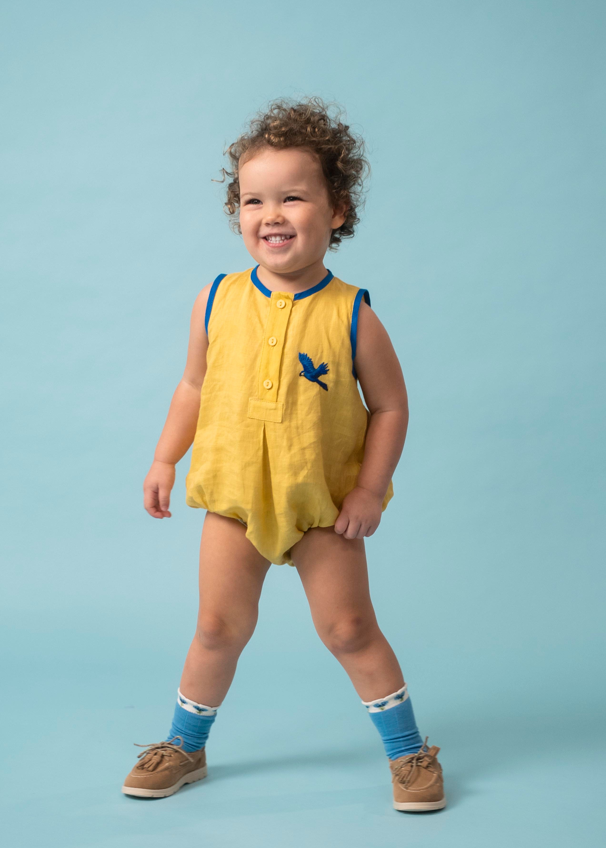 Bebê usando macaquinho infantil amarelo e azul em linho puro com bordado à mão de arara azul