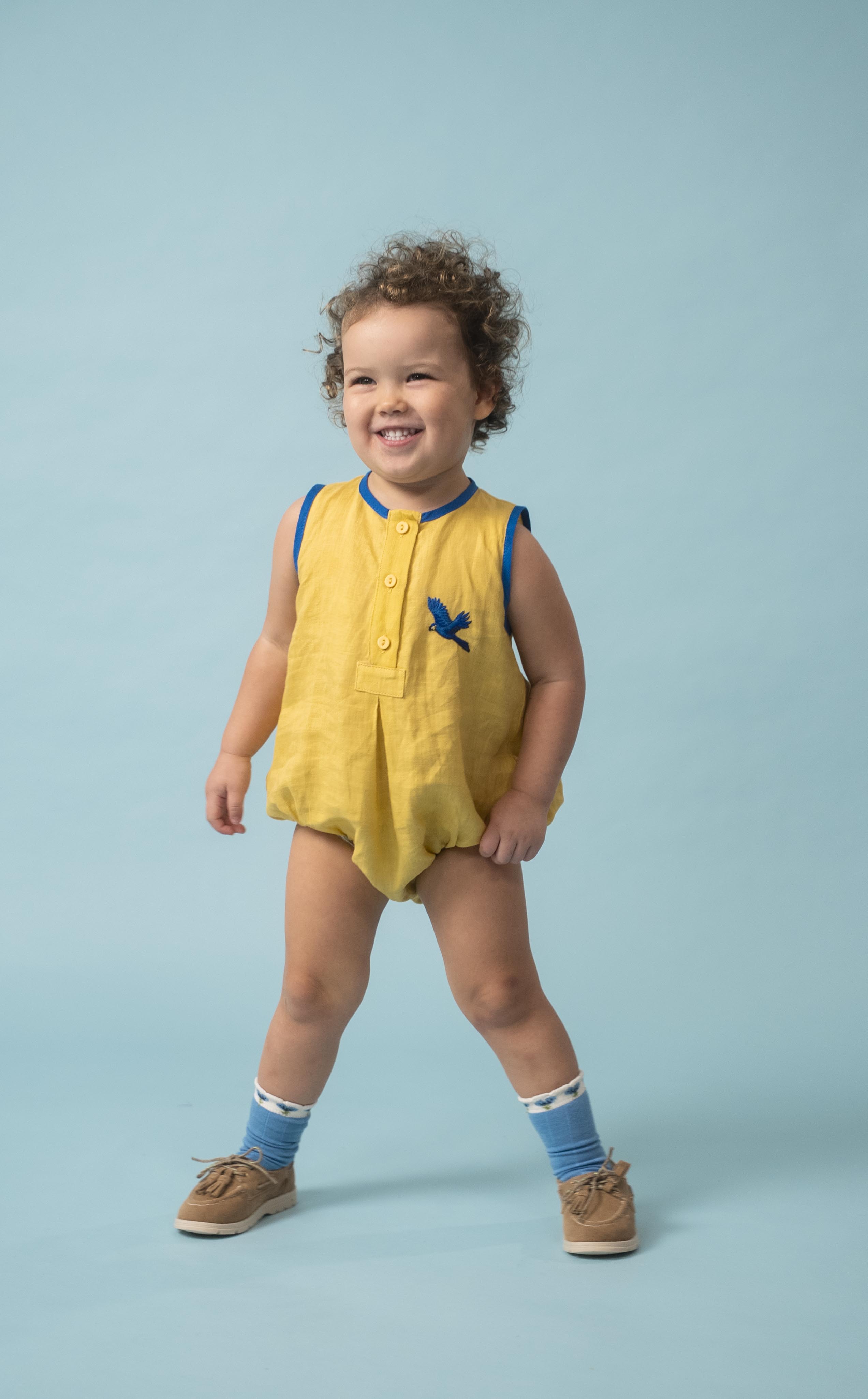 Bebê usando macaquinho infantil amarelo e azul em linho puro com bordado à mão de arara azul