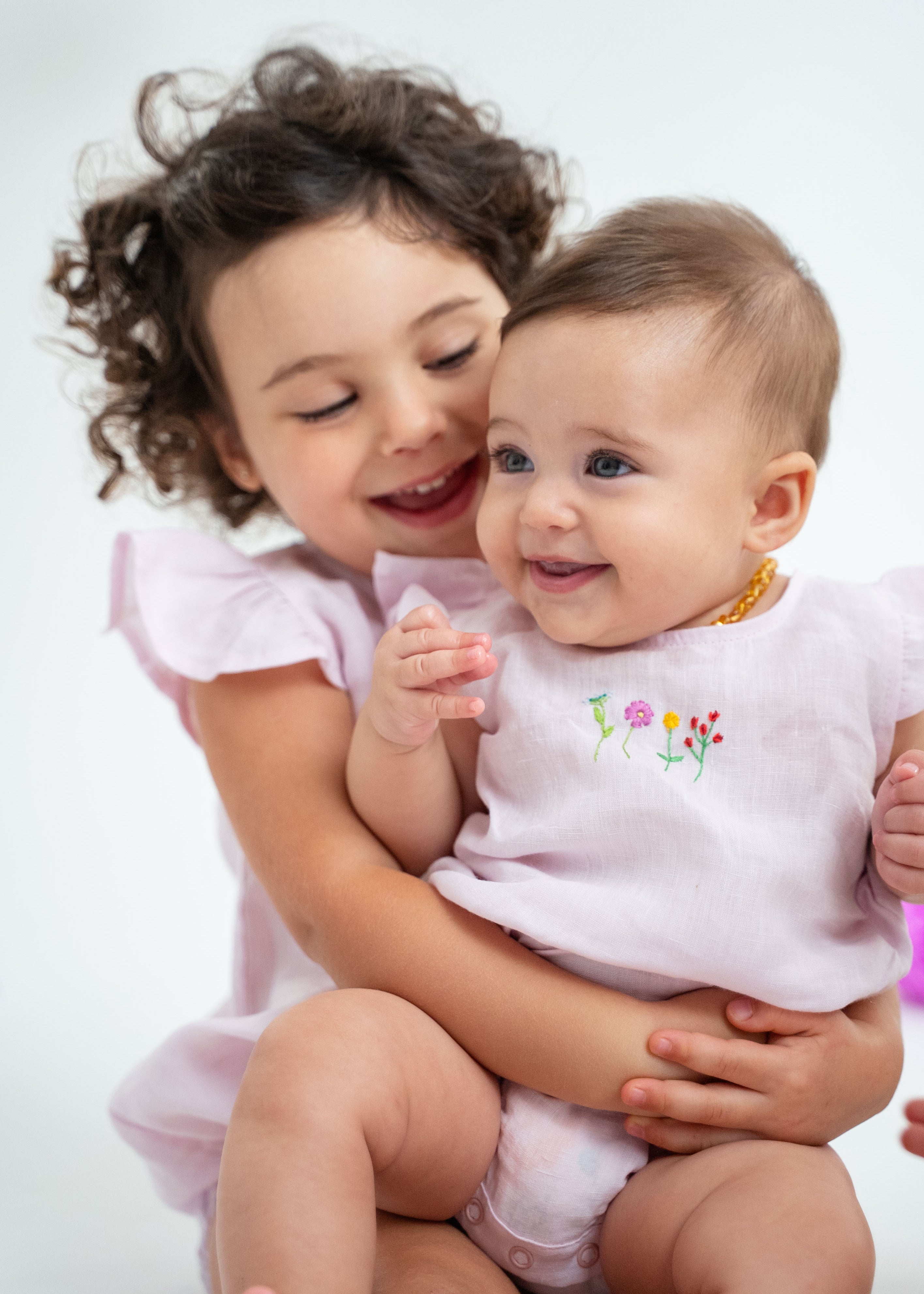 Bebê menina sorrindo no colo da irmã, ambas vestindo macaquinho estilo romper em linho cor de rosa com bordado de flores no peito.