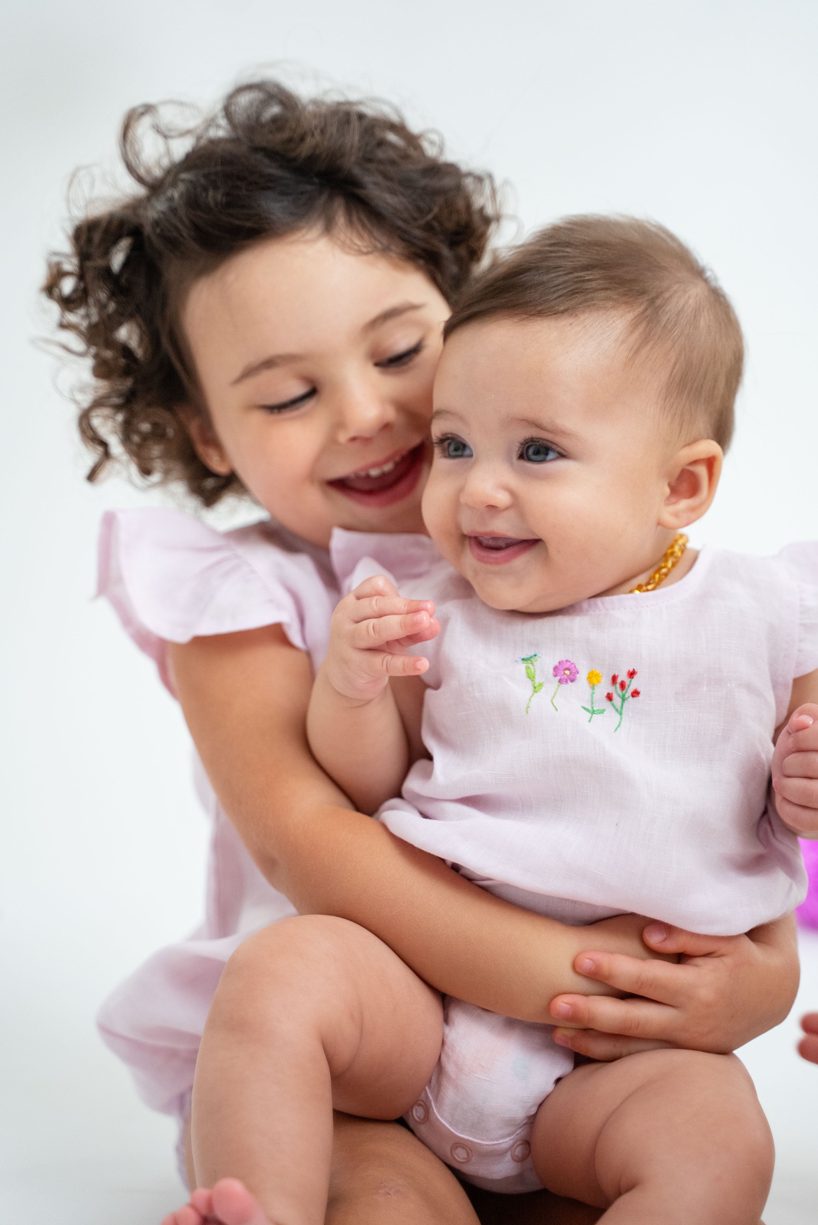 Bebê menina sorrindo no colo da irmã, ambas vestindo macaquinho estilo romper em linho cor de rosa com bordado de flores no peito.
