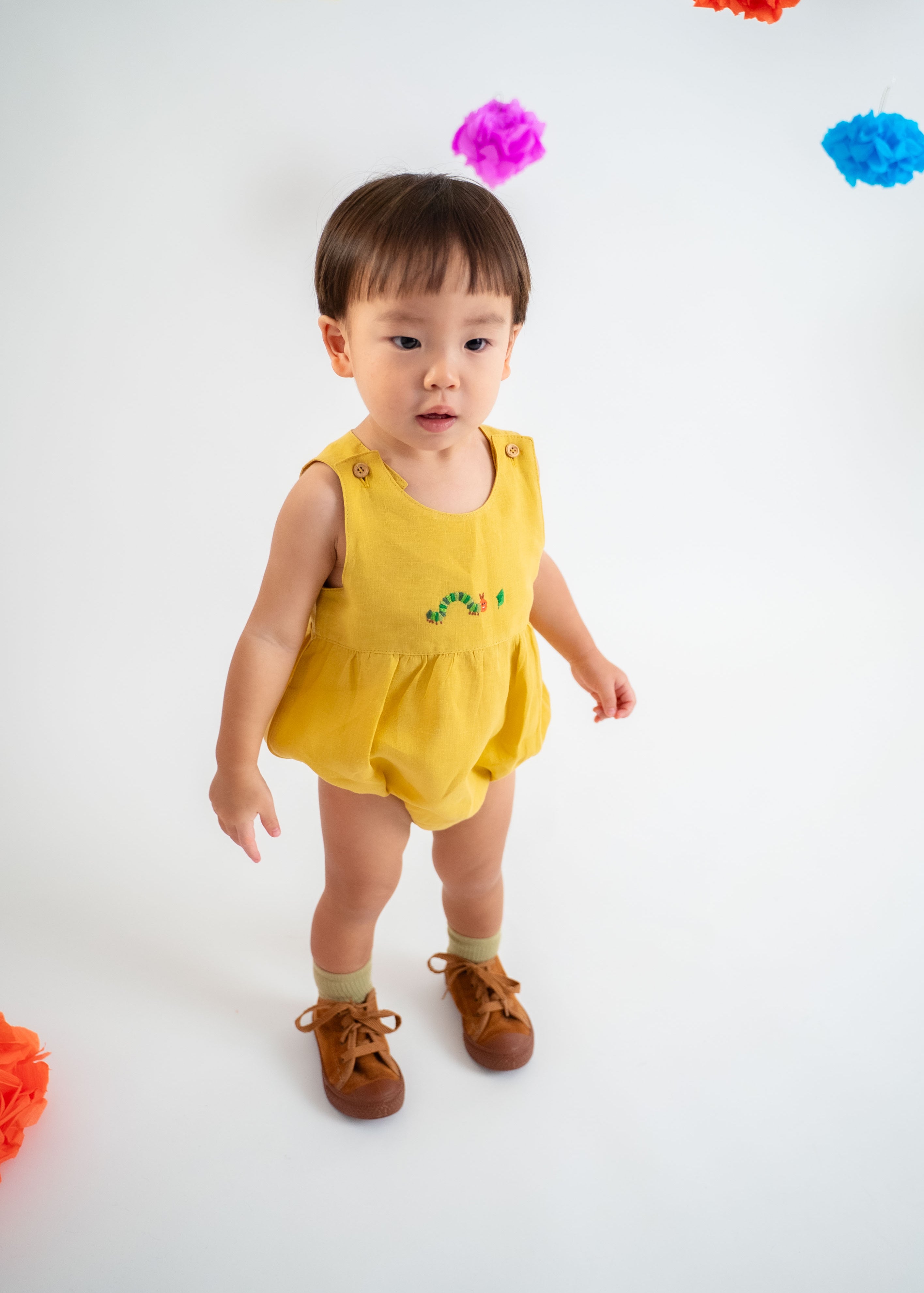 Bebê menino vestindo macaquinho romper unissex em linho amarelo com bordado de lagarta.