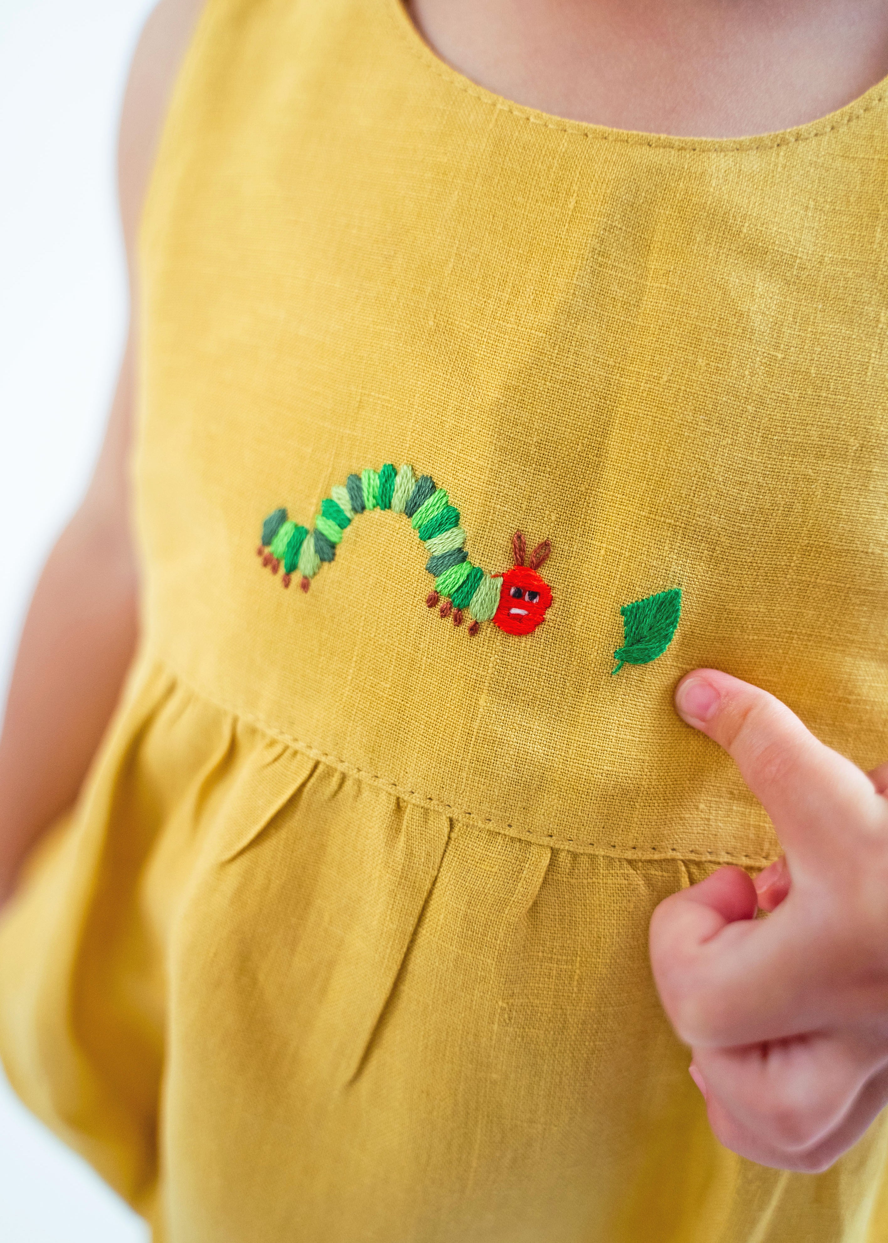 Bebê menino tocando com os dedos o bordado de lagarta do macaquinho unissex estilo romper em linho amarelo.