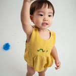 Bebê menino vestindo macaquinho romper unissex em linho amarelo com bordado de lagarta, com uma mão para o alto.