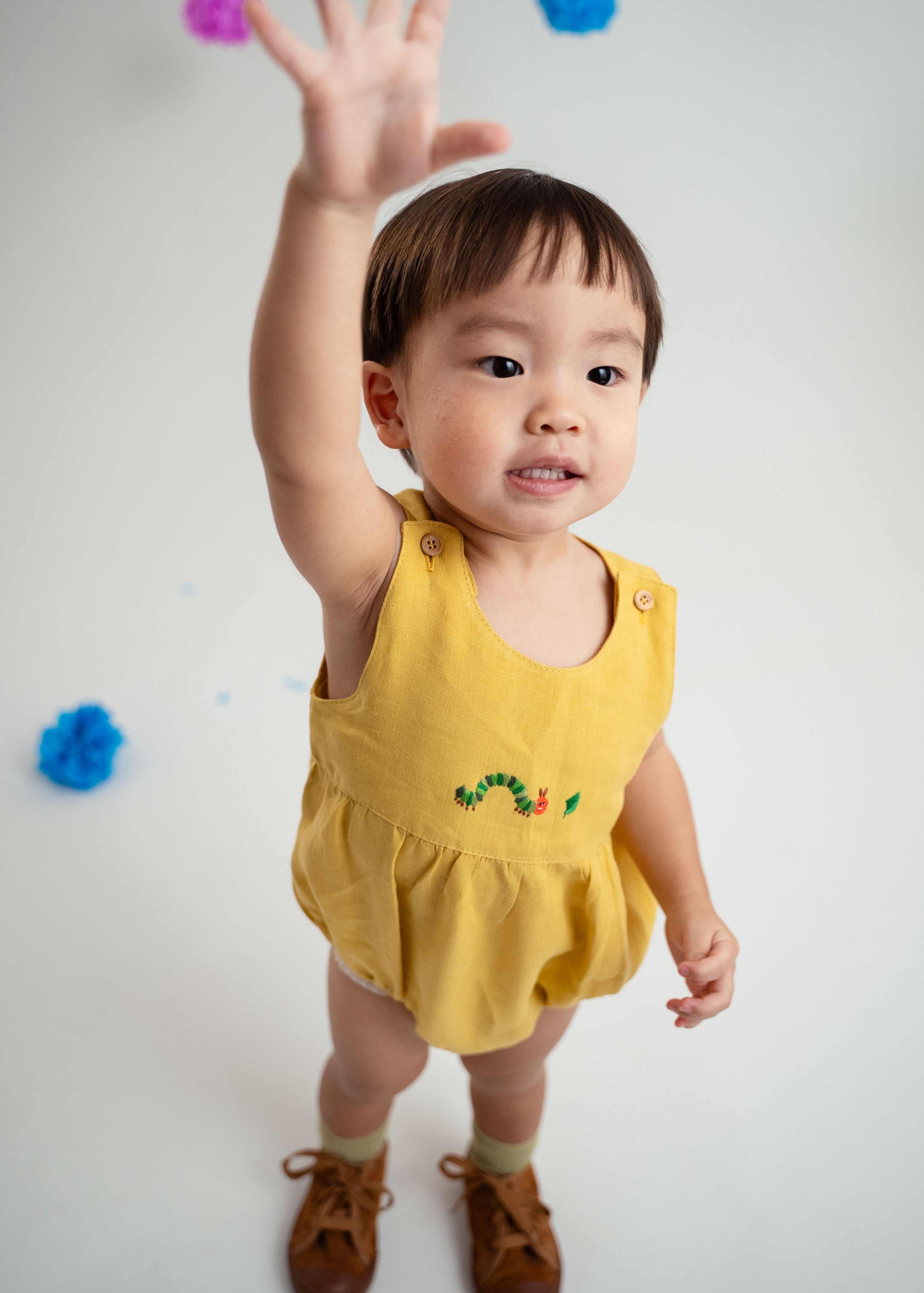 Bebê menino vestindo macaquinho romper unissex em linho amarelo com bordado de lagarta, com uma mão para o alto.