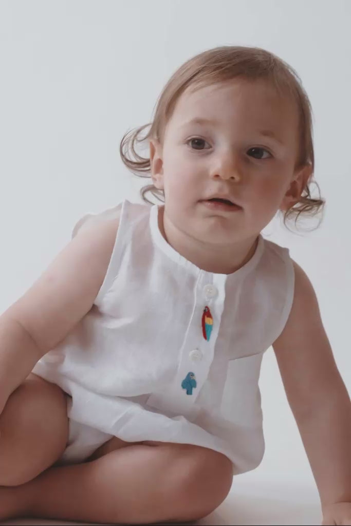 Bebê usando Macaquinho Valen de linho branco com araras bordadas à mão e modelagem soltinha.