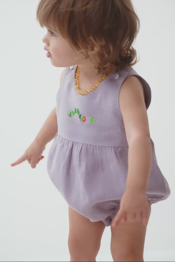 Menina bebê usando macaquinho estilo romper, mostrando o caimento, o tecido de linho puro e o detalhe do bordado de lagarta.
