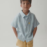 Menino vestindo camisa em linho azul bebê, com close nos detalhes do bordado e acabamento.