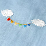 Detalhe do bordado à mão do Conjunto Duda, com nuvens brancas em relevo e bandeirinhas coloridas sobre linho azul celeste.