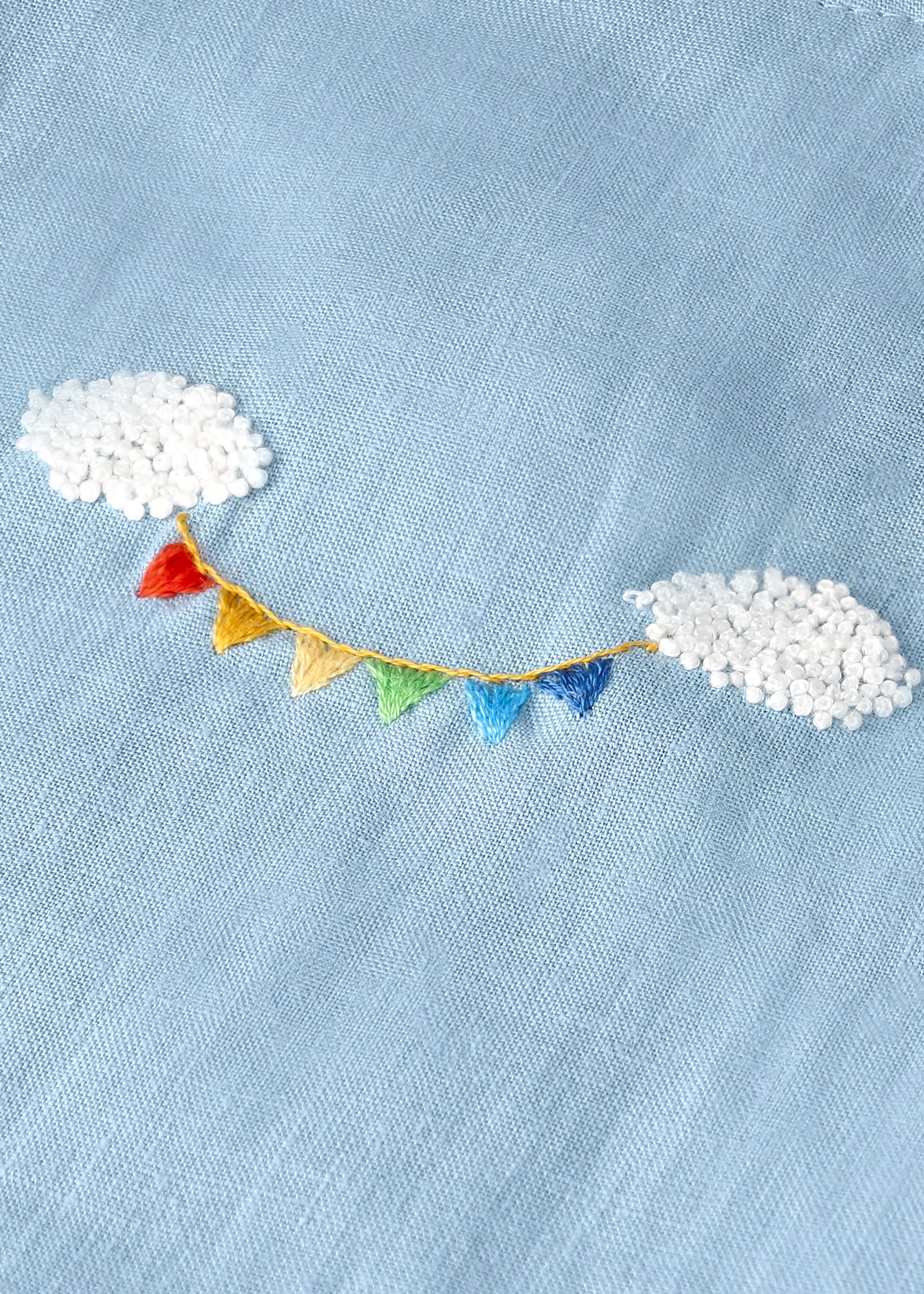 Detalhe do bordado à mão do Conjunto Duda, com nuvens brancas em relevo e bandeirinhas coloridas sobre linho azul celeste.