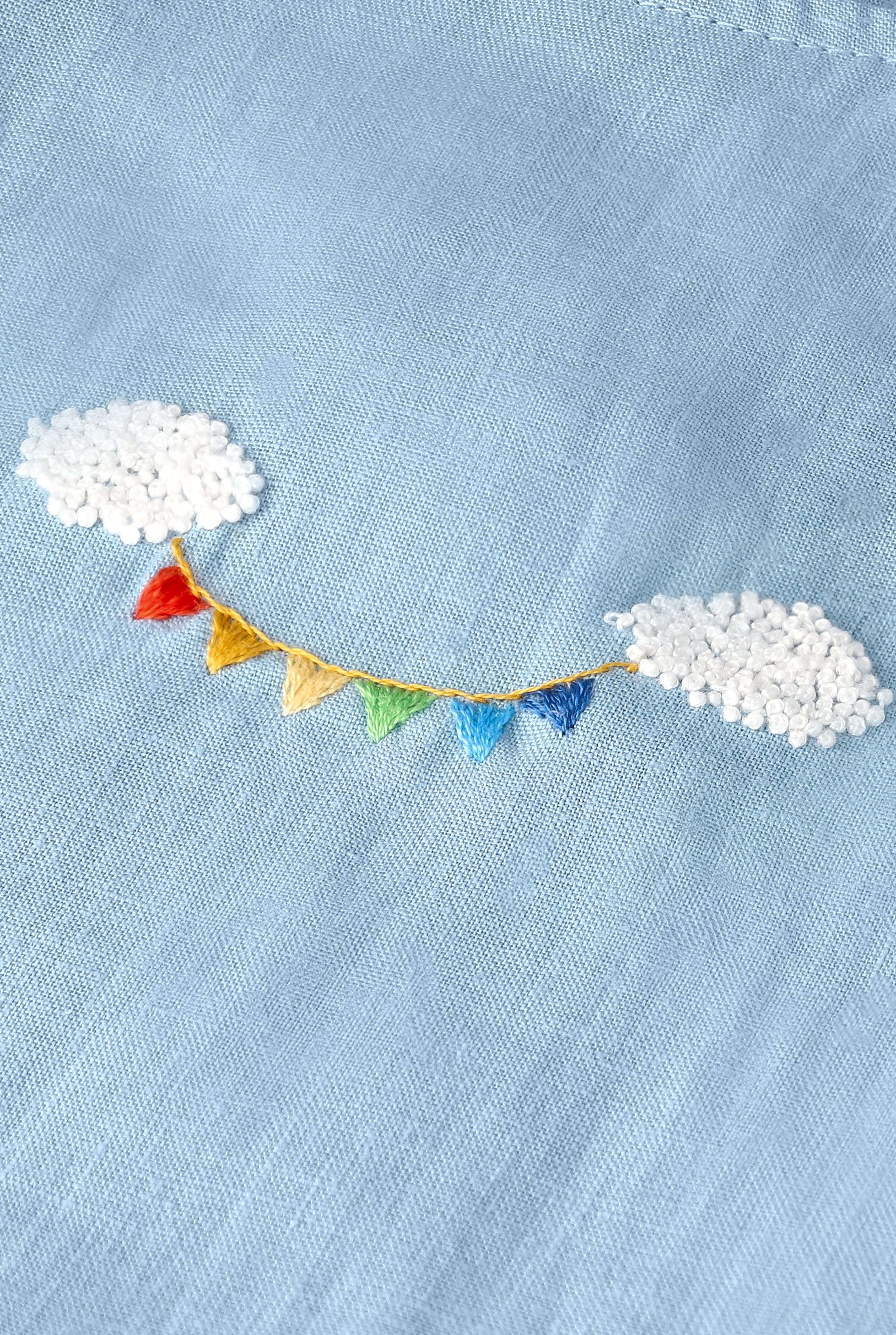 Detalhe do bordado à mão do Conjunto Duda, com nuvens brancas em relevo e bandeirinhas coloridas sobre linho azul celeste.