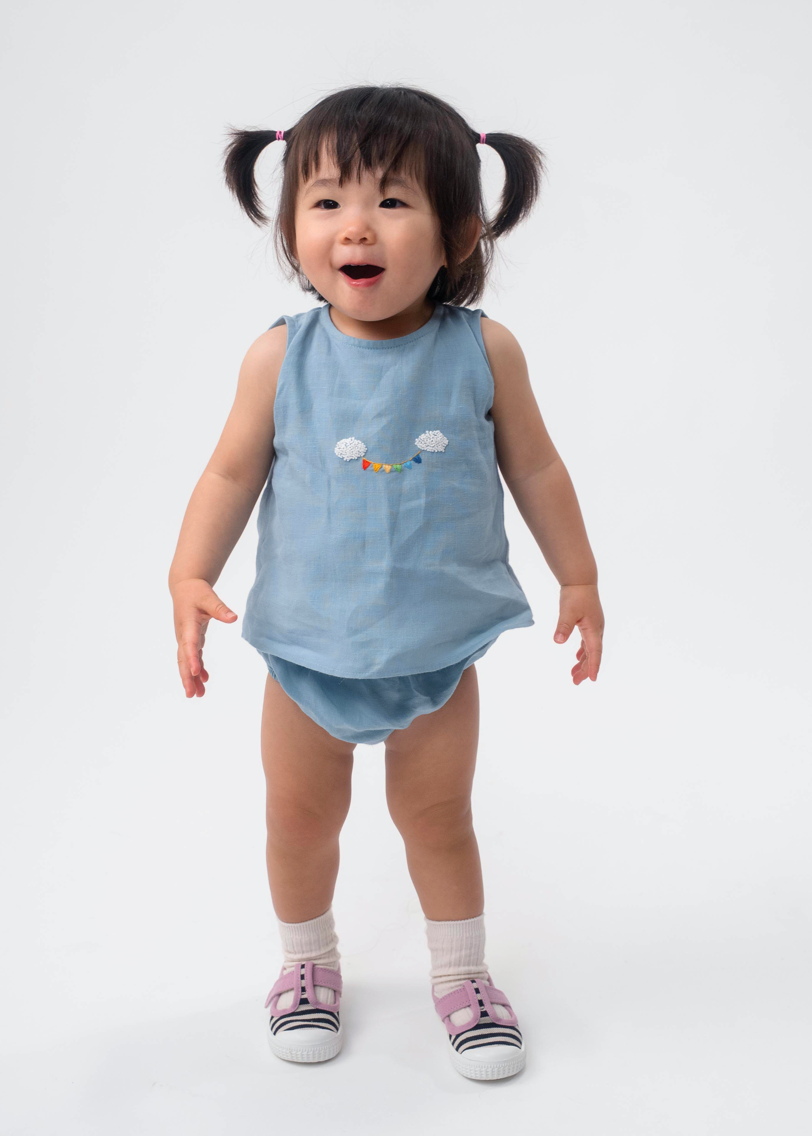 Bebê vestindo Conjunto Duda em linho azul celeste, com regata bordada à mão e tapa-fralda.