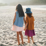 Duas meninas de mãos dadas de costas na praia, uma vestindo saia infantil roxa e outra saia em linho listrado rosa, ambas com camisas em linho colorido.