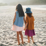 Duas meninas de mãos dadas de costas na praia, uma vestindo saia infantil roxa e outra saia em linho listrado rosa, ambas com camisas em linho colorido.