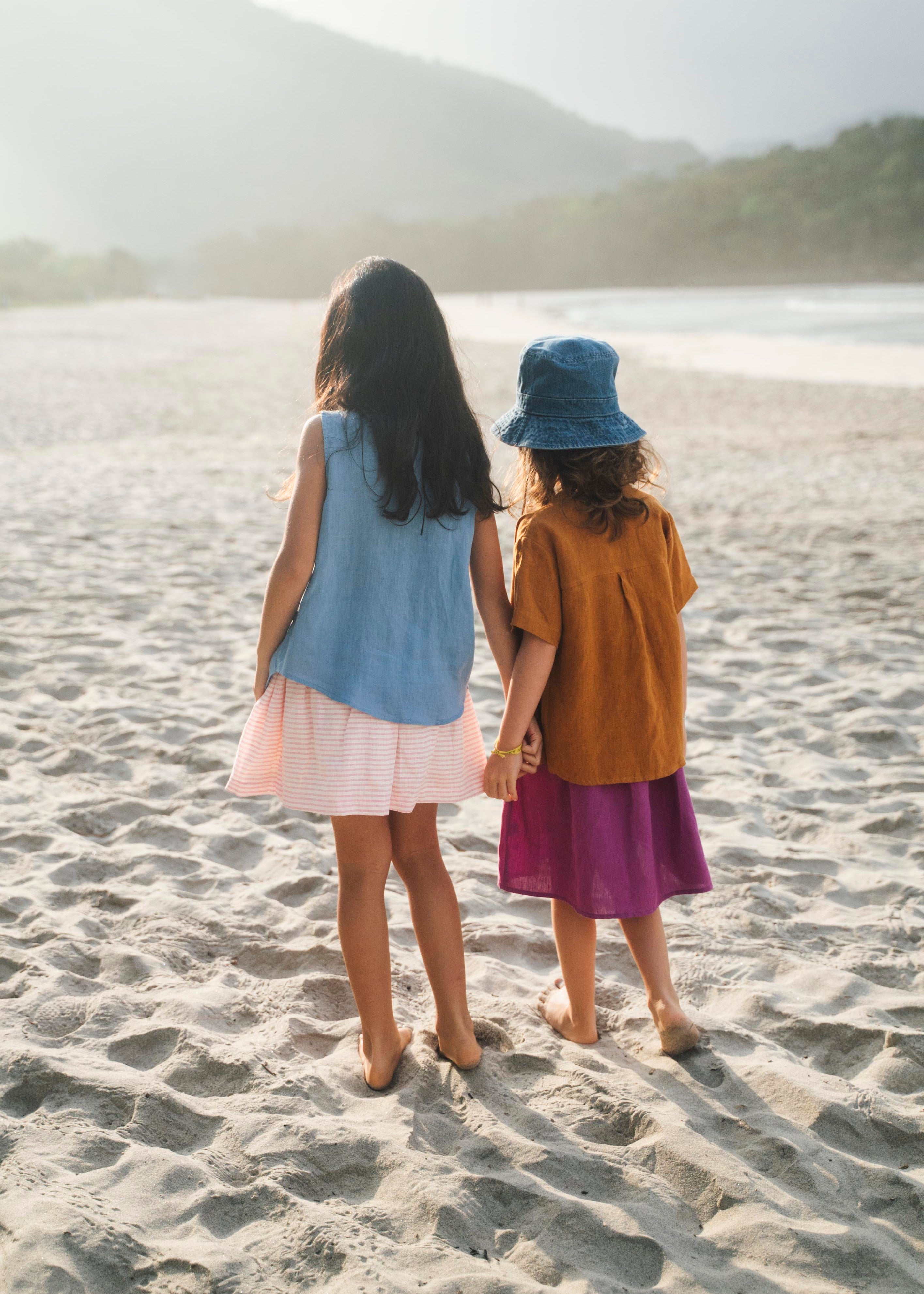 Duas meninas de mãos dadas de costas na praia, uma vestindo saia infantil roxa e outra saia em linho listrado rosa, ambas com camisas em linho colorido.