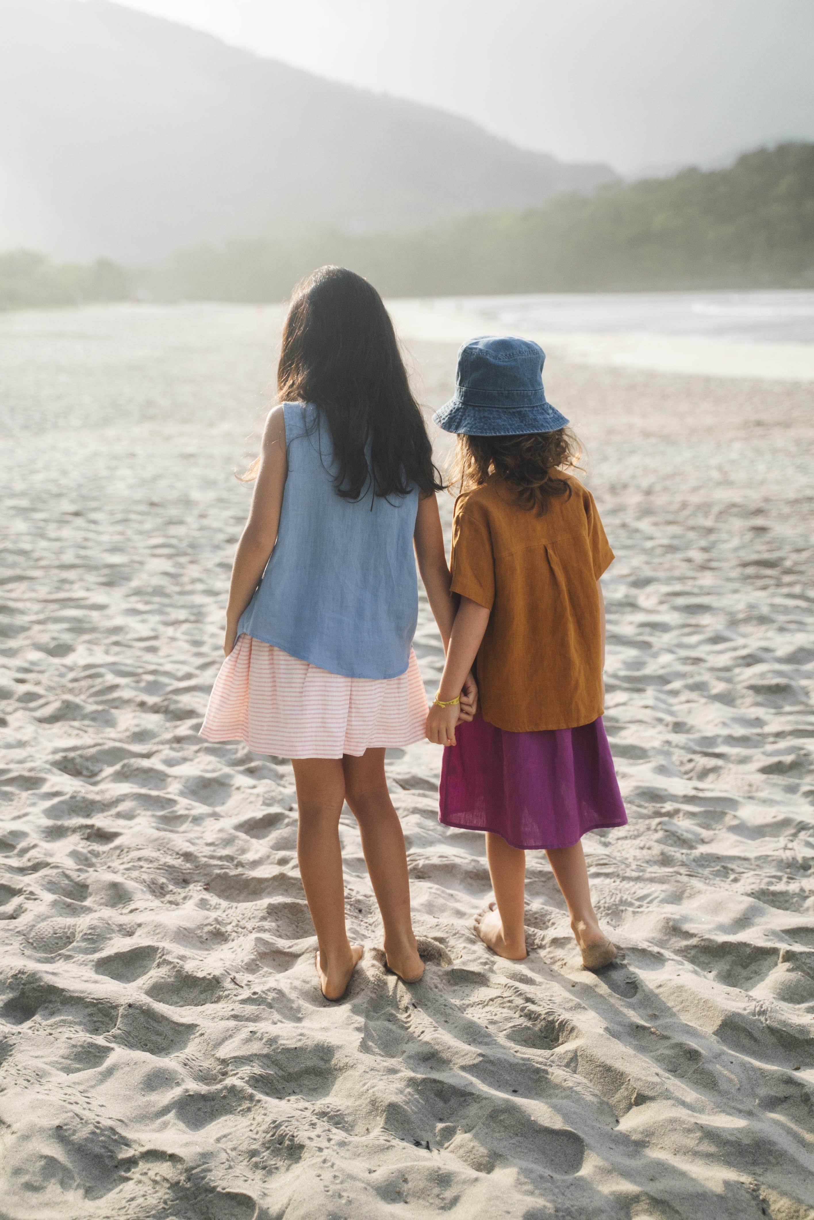 Duas meninas de mãos dadas de costas na praia, uma vestindo saia infantil roxa e outra saia em linho listrado rosa, ambas com camisas em linho colorido.