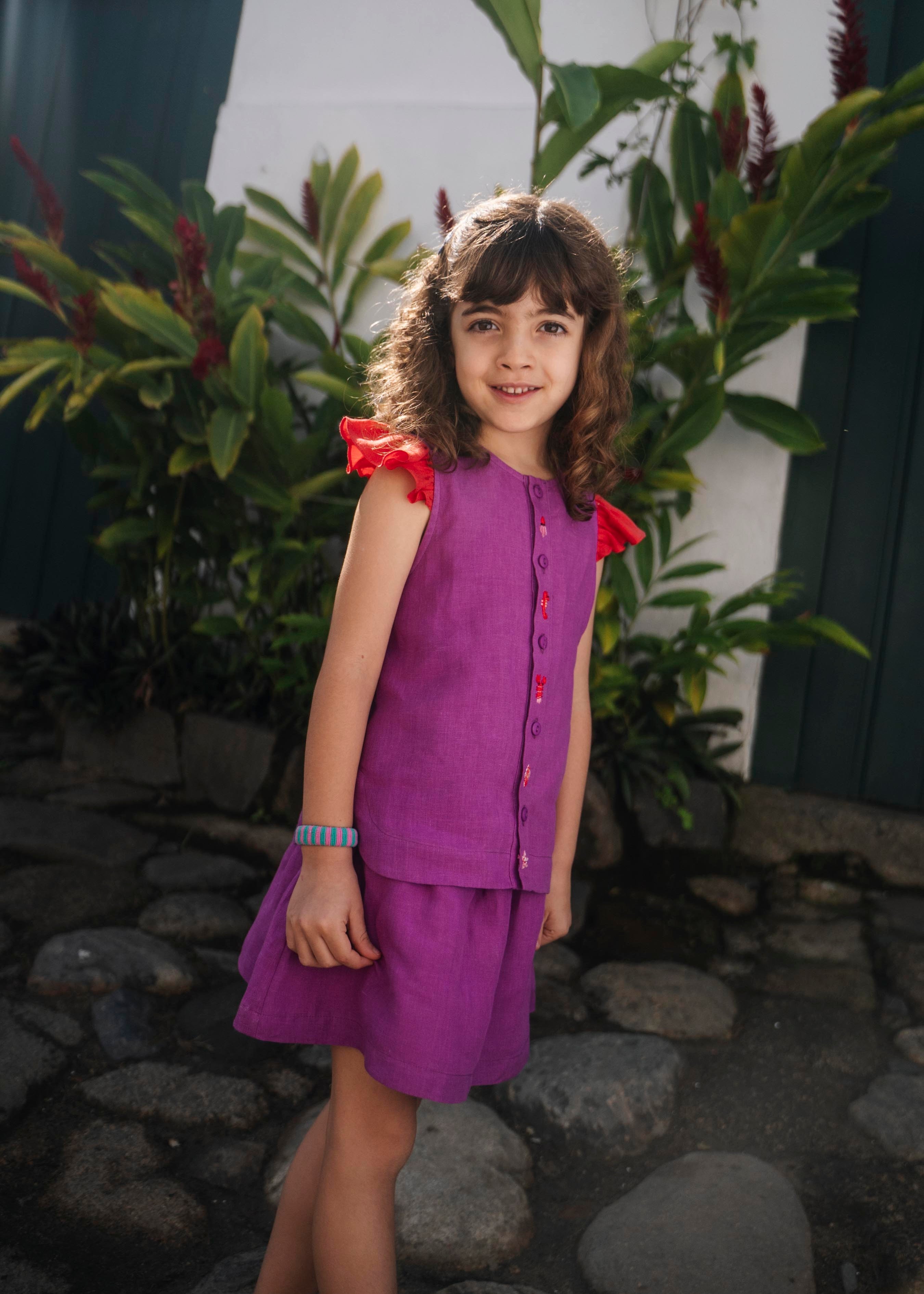 Menina usando saia infantil em linho roxo combinada com camisa da mesma cor, em ambiente externo.
