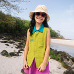 Menina na praia vestindo saia infantil em linho roxo combinada com camisa verde sem mangas, criando contraste divertido.