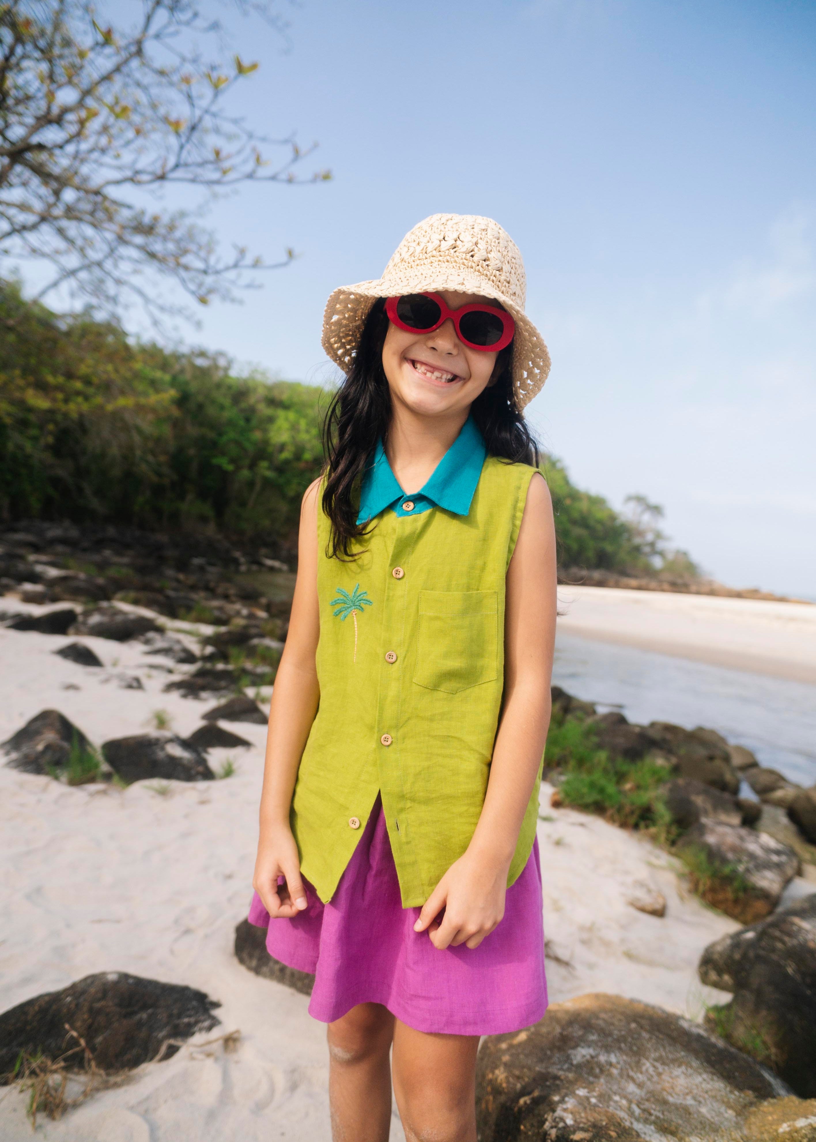 Menina na praia vestindo saia infantil em linho roxo combinada com camisa verde sem mangas, criando contraste divertido.