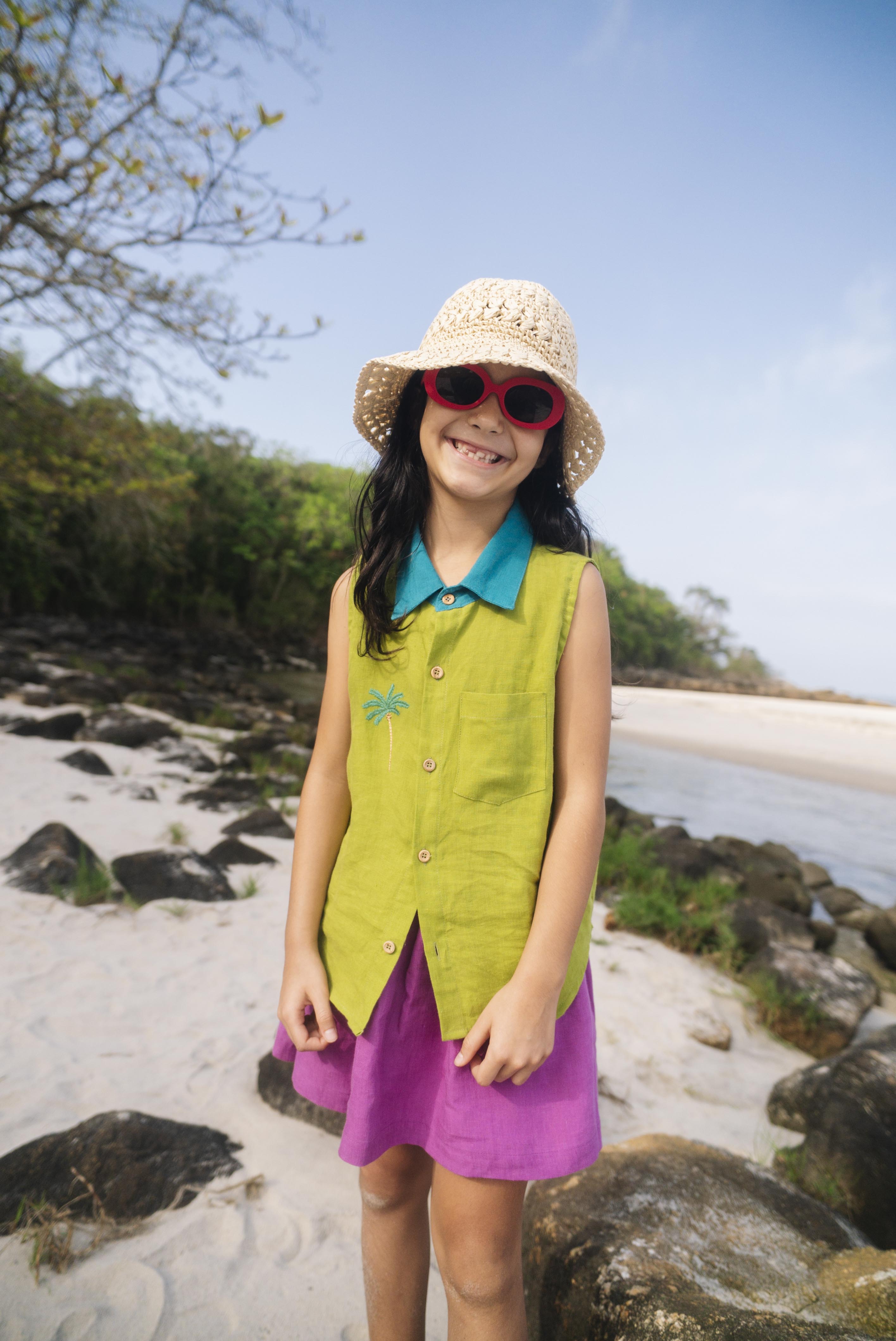 Menina na praia vestindo saia infantil em linho roxo combinada com camisa verde sem mangas, criando contraste divertido.