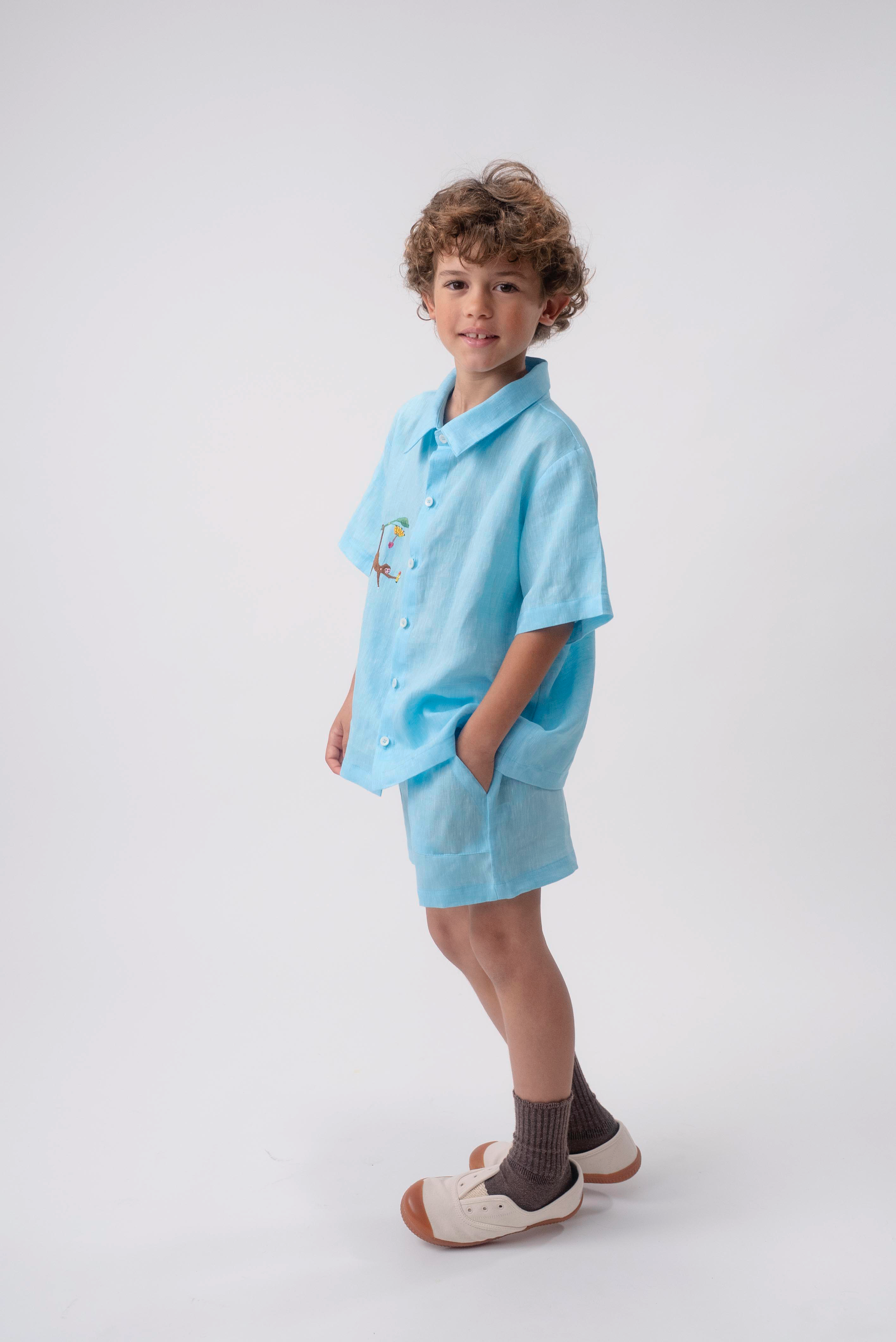 Menino de perfil vestindo Shorts Chico infantil em linho azul turquesa, com destaque para o bolso frontal e a modelagem soltinha.