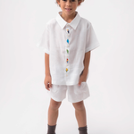 Menino vestindo Shorts Chico infantil em linho branco, combinado com camisa branca bordada, em fundo claro.
