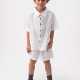 Menino vestindo Shorts Chico infantil em linho branco, combinado com camisa branca bordada, em fundo claro.