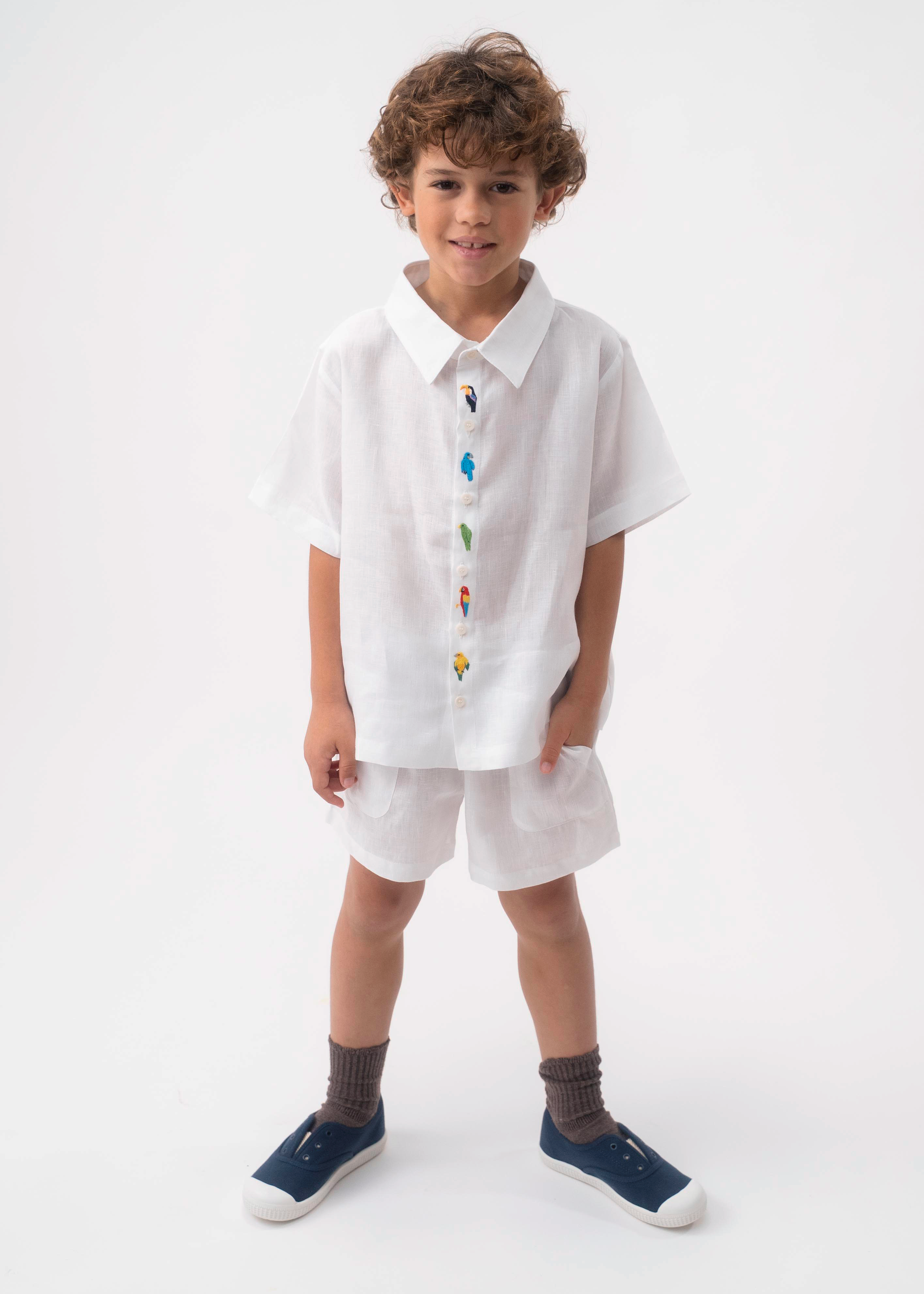 Menino vestindo Shorts Chico infantil em linho branco, combinado com camisa branca bordada, em fundo claro.