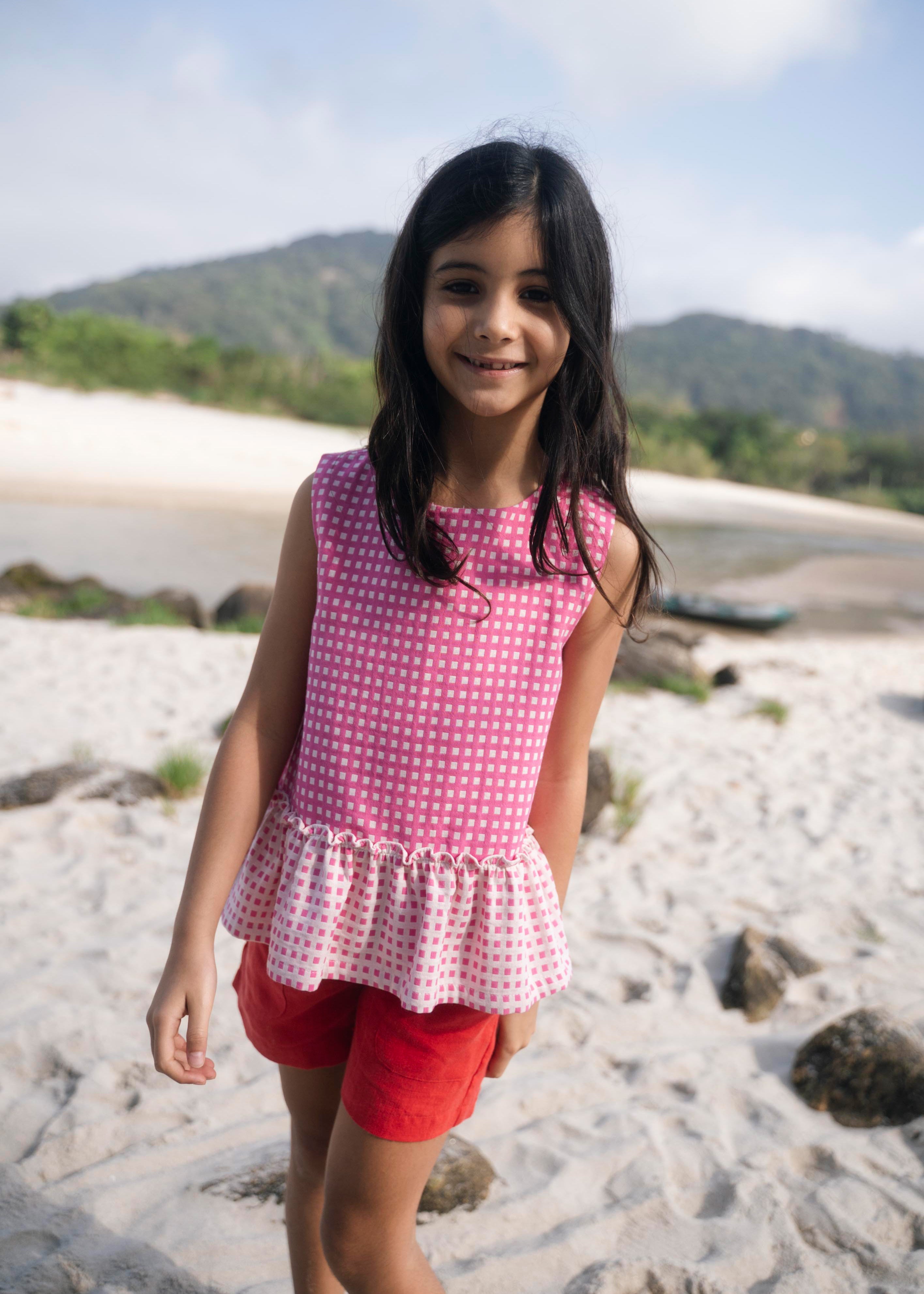 Menina sorrindo na praia usando shorts infantil em linho puro vermelho com bata vichy rosa sem mangas e com babado.