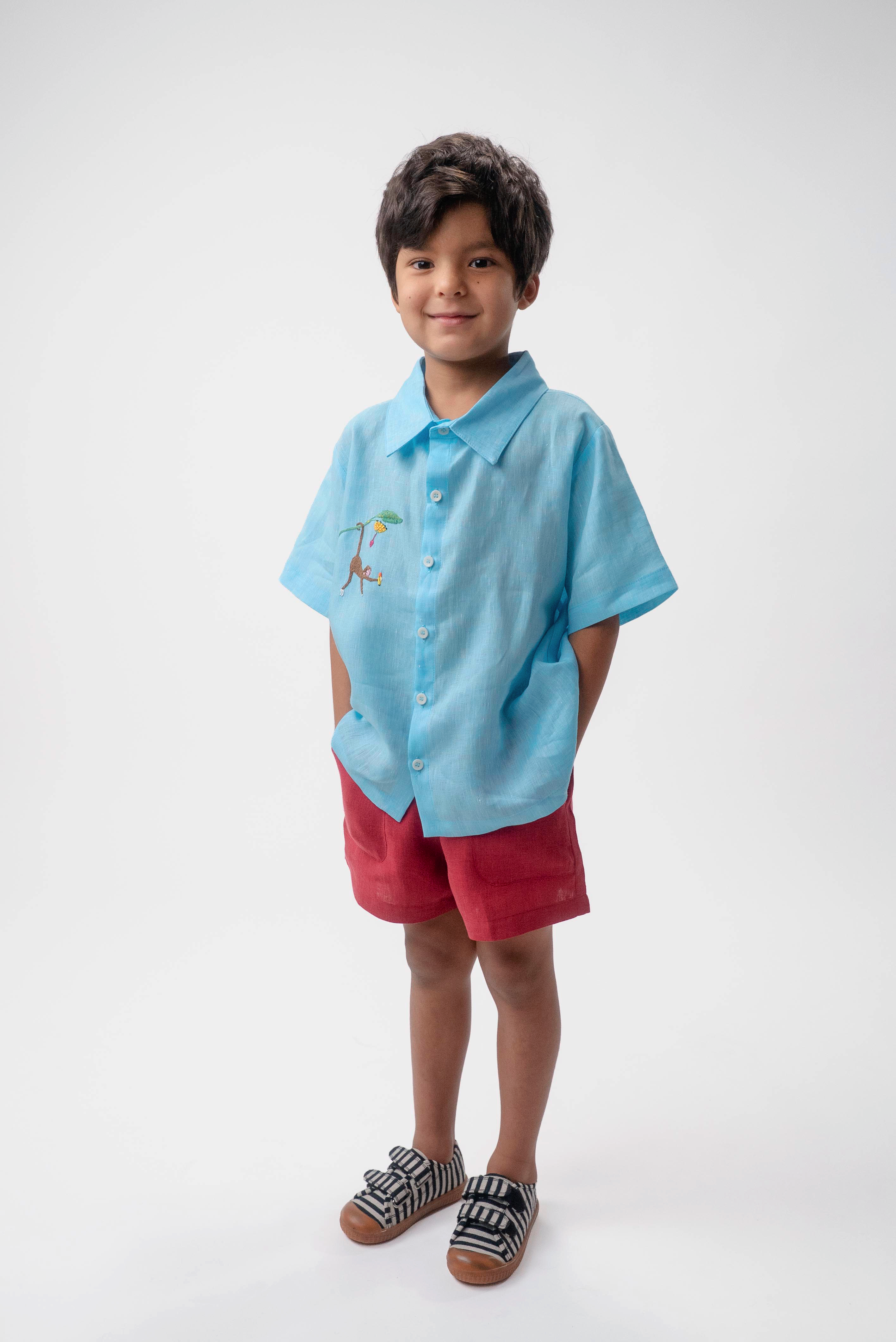 Menino vestindo Shorts Chico infantil em linho vinho com camisa azul, modelo de shorts para menino e menina em fundo branco.