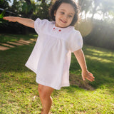 Menina usando vestido bebê em algodão branco e rosa listrado com gola xadrez bordada de morangos, em dia de sol.