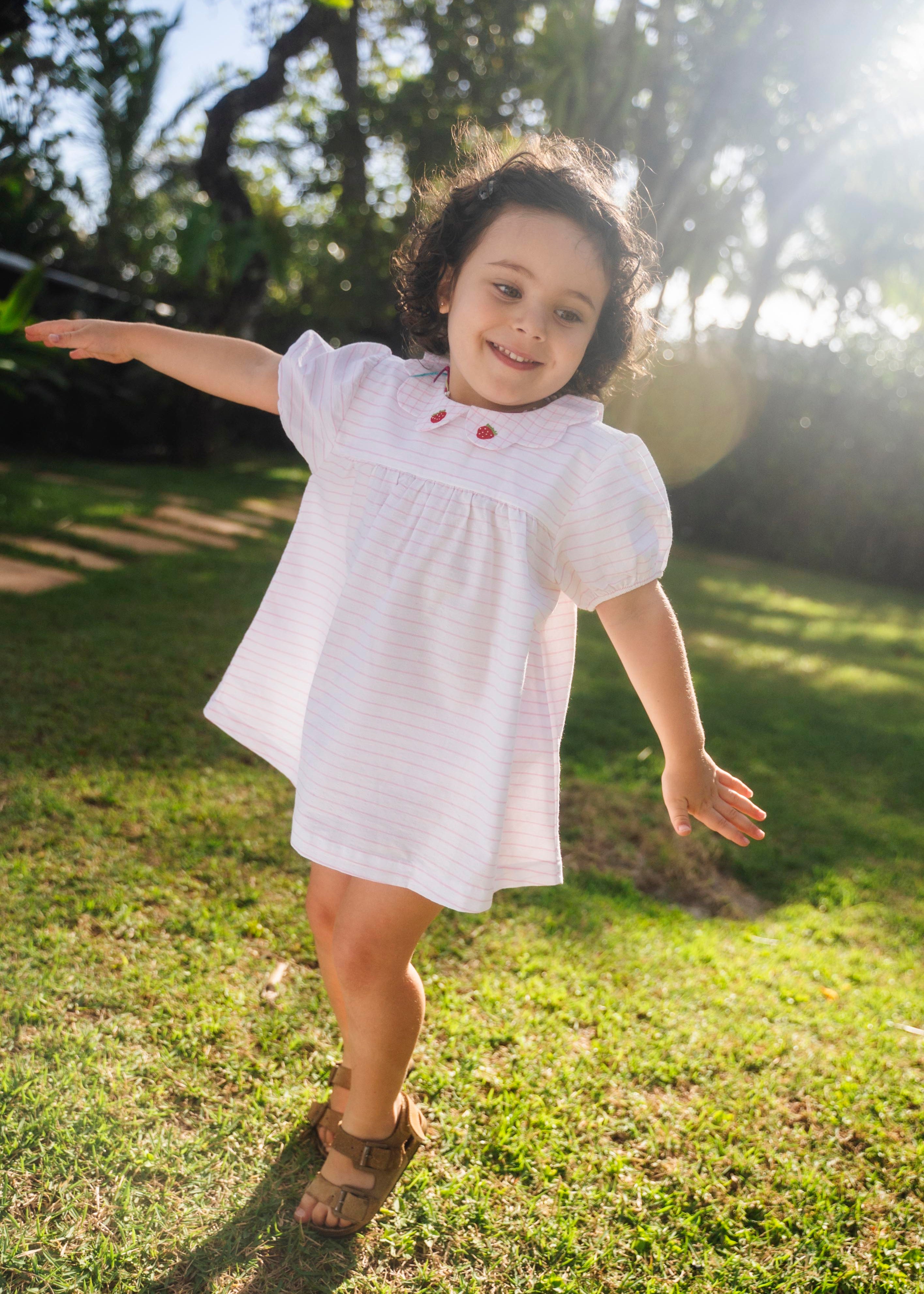 Menina usando vestido bebê em algodão branco e rosa listrado com gola xadrez bordada de morangos, em dia de sol.