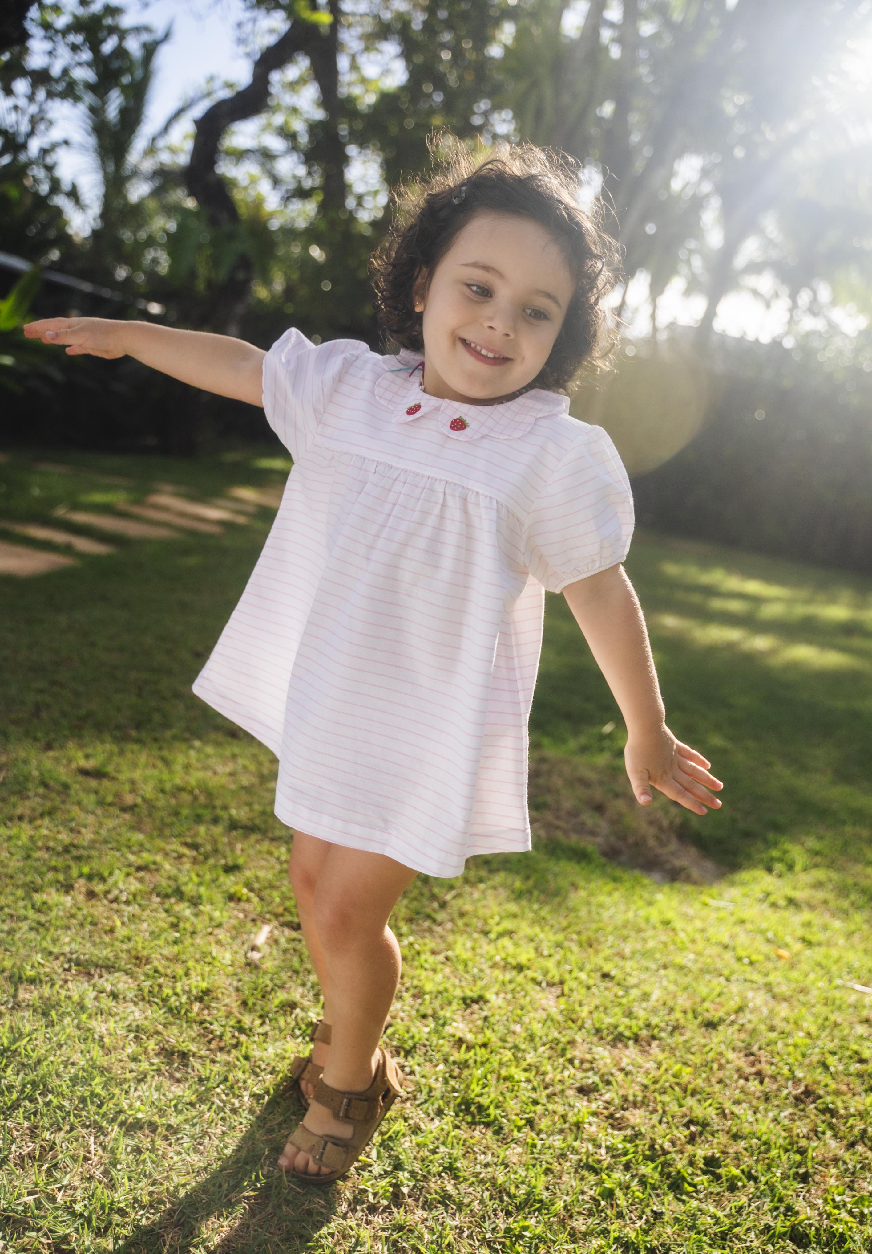 Menina usando vestido bebê em algodão branco e rosa listrado com gola xadrez bordada de morangos, em dia de sol.