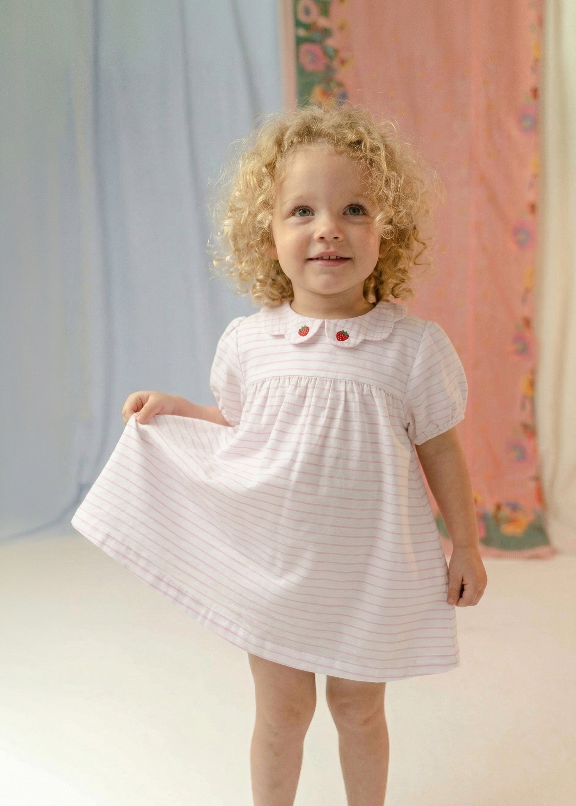 Menina usando vestido bebê em algodão branco e rosa listrado com gola xadrez bordada de morangos.