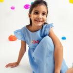 Menina sentada no chão com vestido infantil em linho azul-celeste com bordado de flores e mangas drapeadas