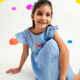 Menina sentada no chão com vestido infantil em linho azul-celeste com bordado de flores e mangas drapeadas