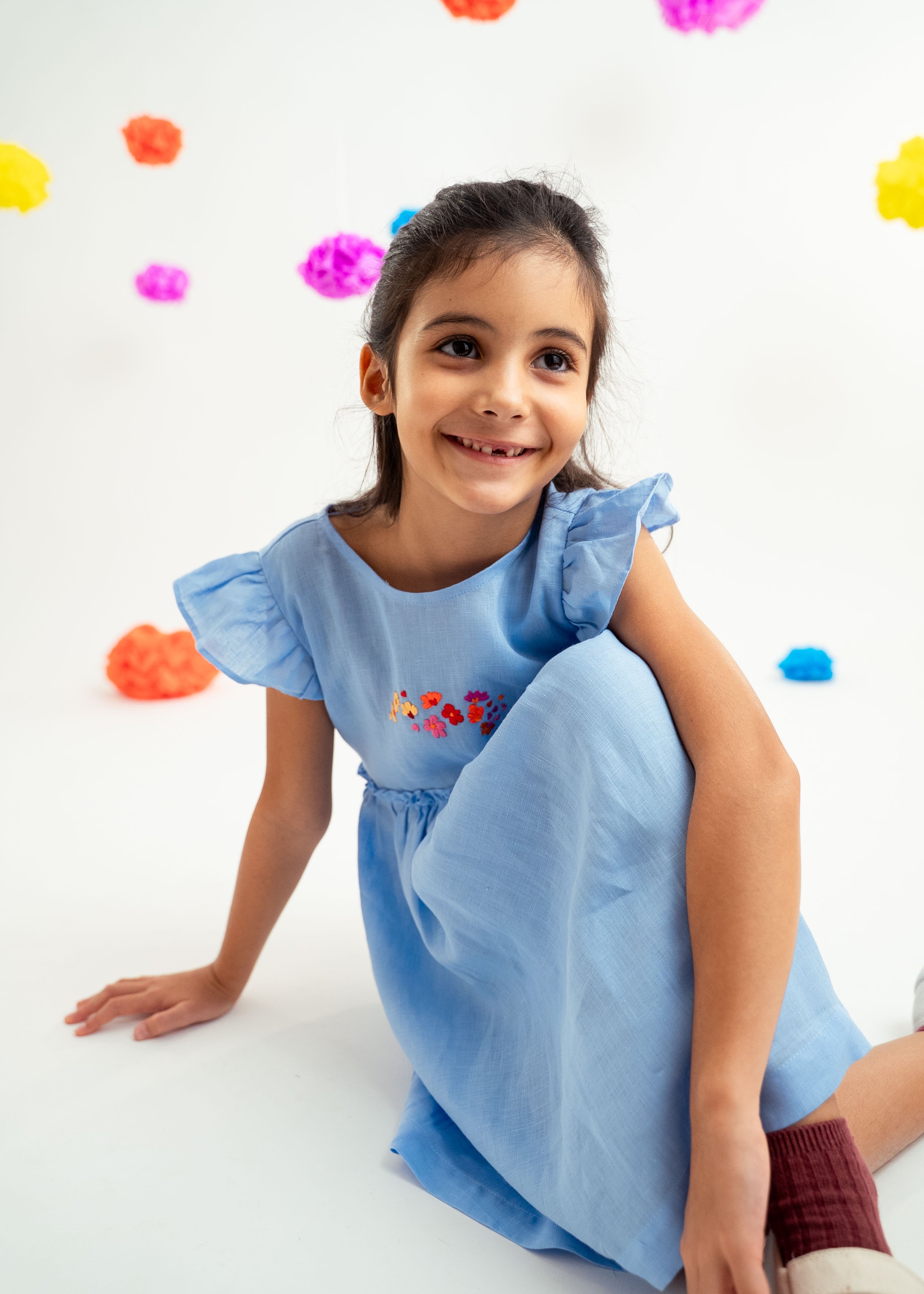 Menina sentada no chão com vestido infantil em linho azul-celeste com bordado de flores e mangas drapeadas