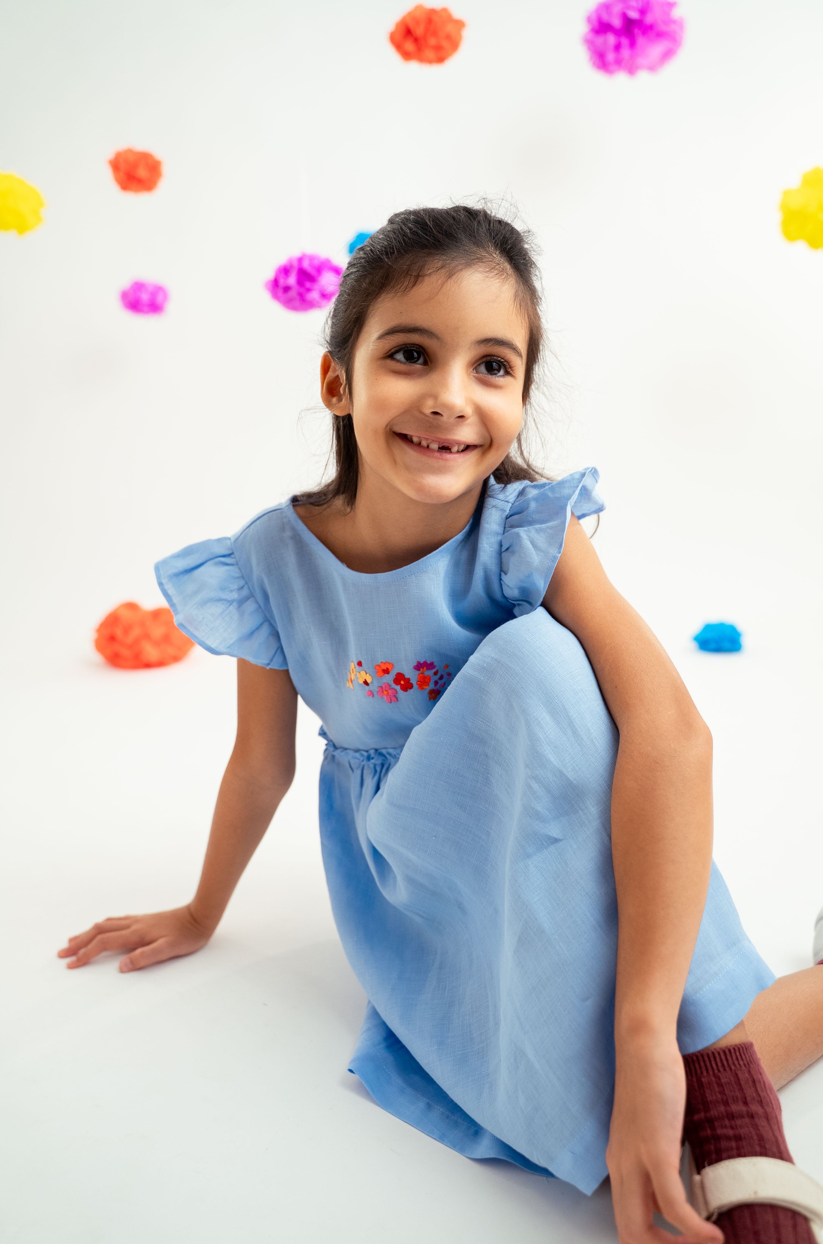 Menina sentada no chão com vestido infantil em linho azul-celeste com bordado de flores e mangas drapeadas