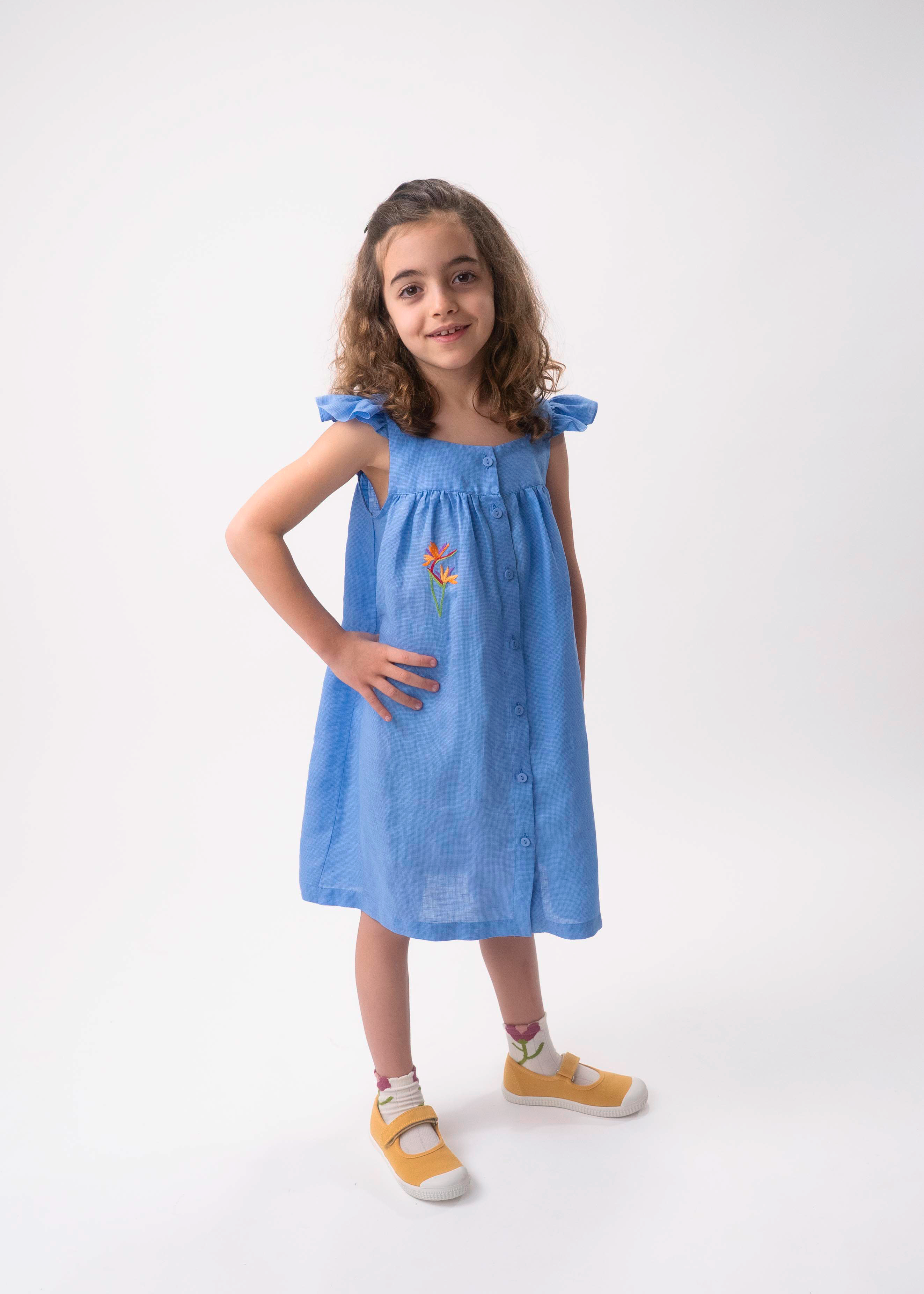 Menina vestindo Vestido Clara infantil em linho azul índigo com helicônia bordada à mão e mangas em babado.