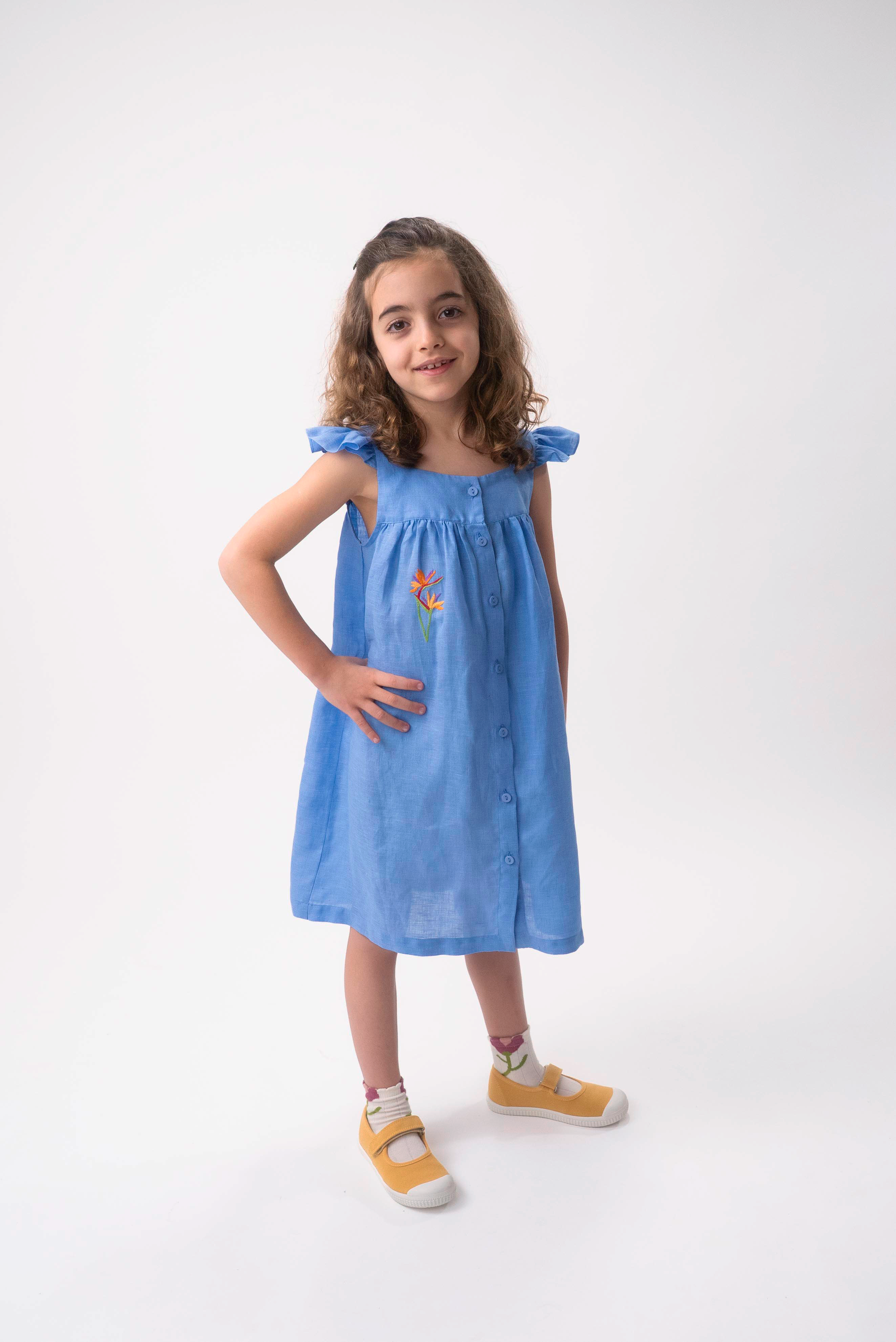 Menina vestindo Vestido Clara infantil em linho azul índigo com helicônia bordada à mão e mangas em babado.