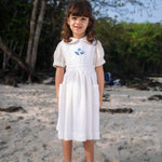 Menina usando vestido infantil de linho branco com bolsos e bordado azul de conchas do mar na pala, em cenário de praia.