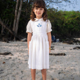 Menina usando vestido infantil de linho branco com bolsos e bordado azul de conchas do mar na pala, em cenário de praia.