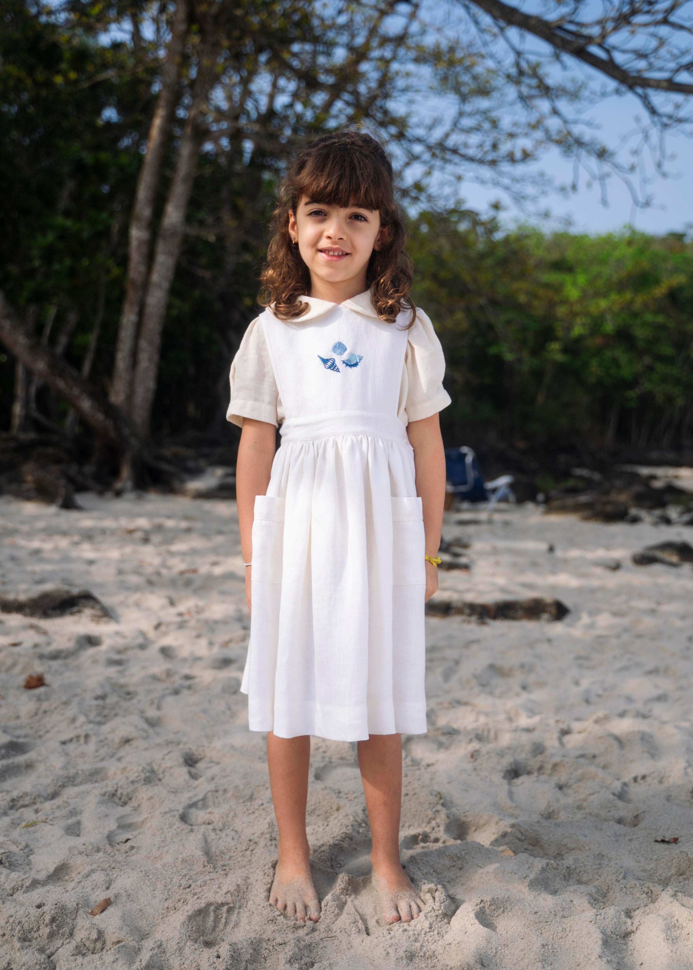 Menina usando vestido infantil de linho branco com bolsos e bordado azul de conchas do mar na pala, em cenário de praia.