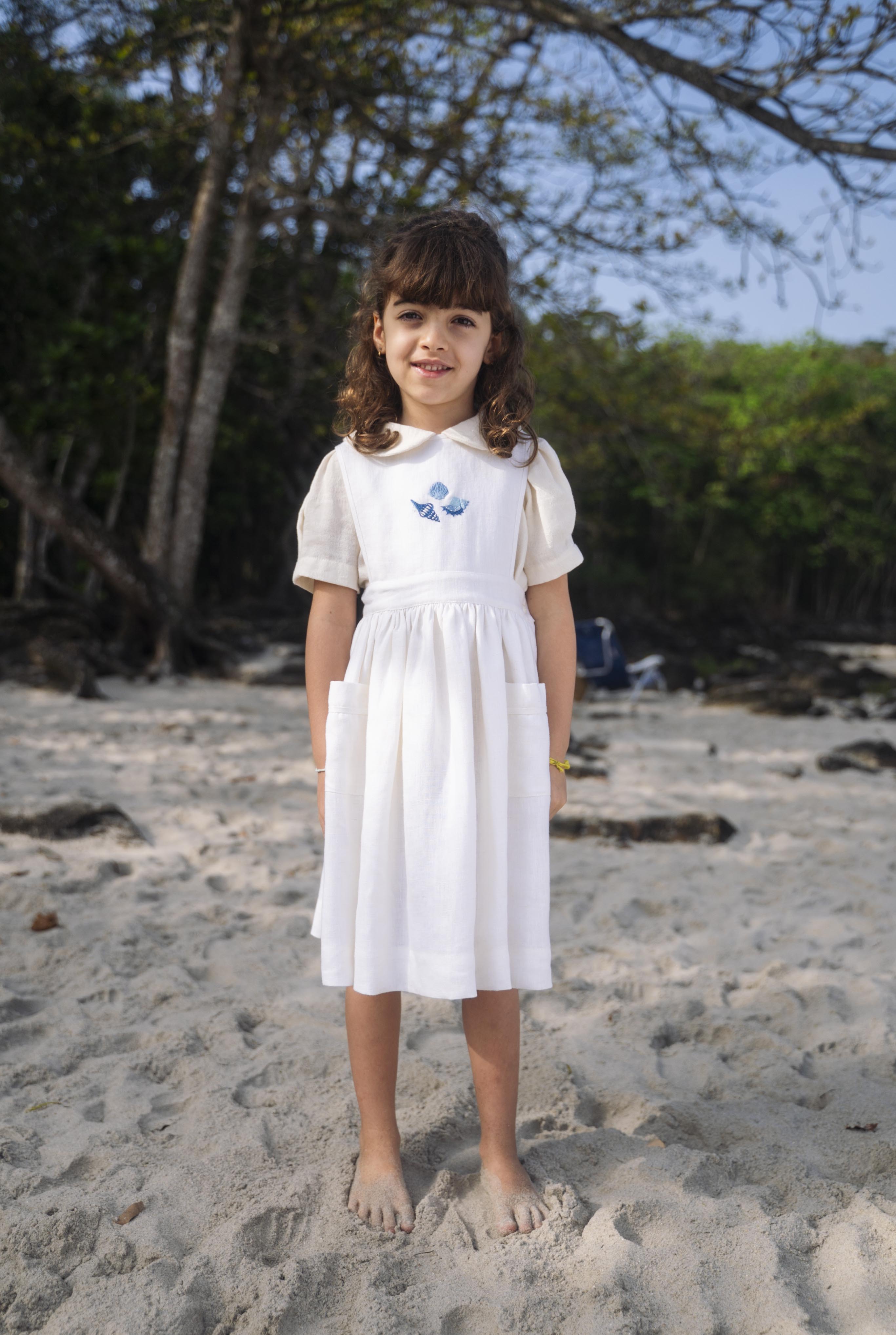 Menina usando vestido infantil de linho branco com bolsos e bordado azul de conchas do mar na pala, em cenário de praia.