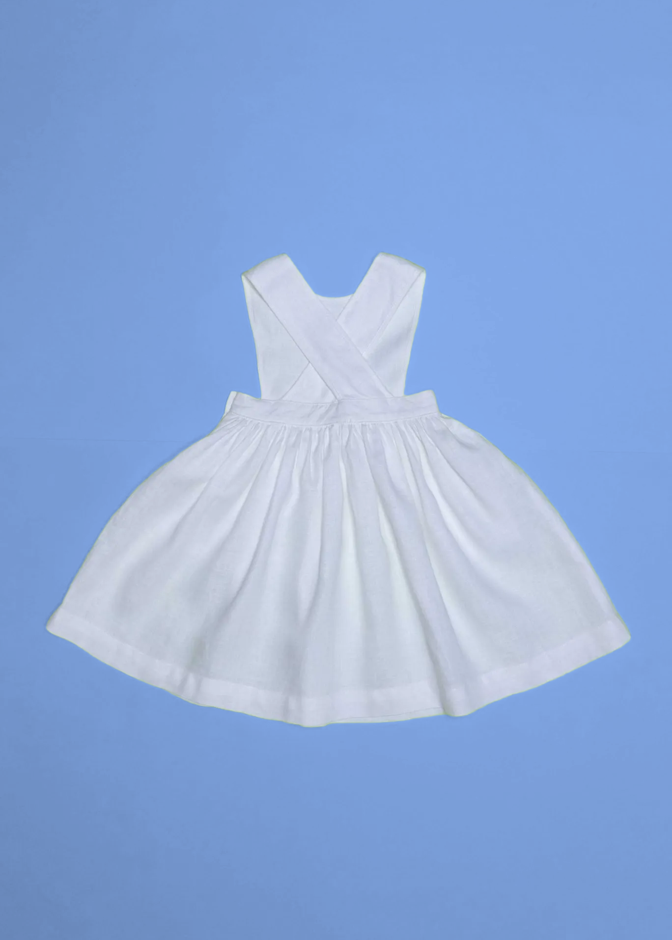 Vista de costas do vestido infantil de linho branco com alças cruzadas e saia ampla, em superfície azul.