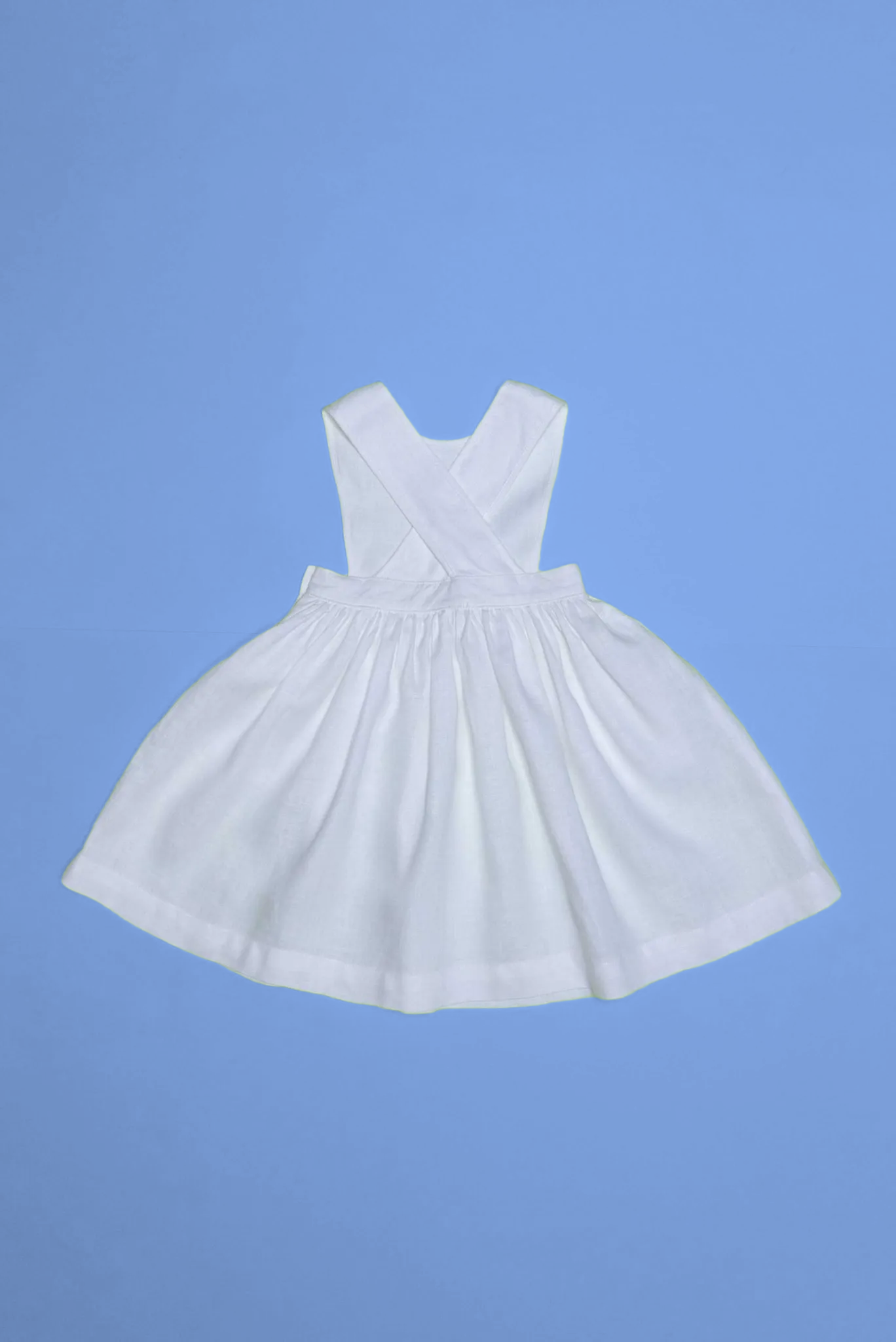 Vista de costas do vestido infantil de linho branco com alças cruzadas e saia ampla, em superfície azul.