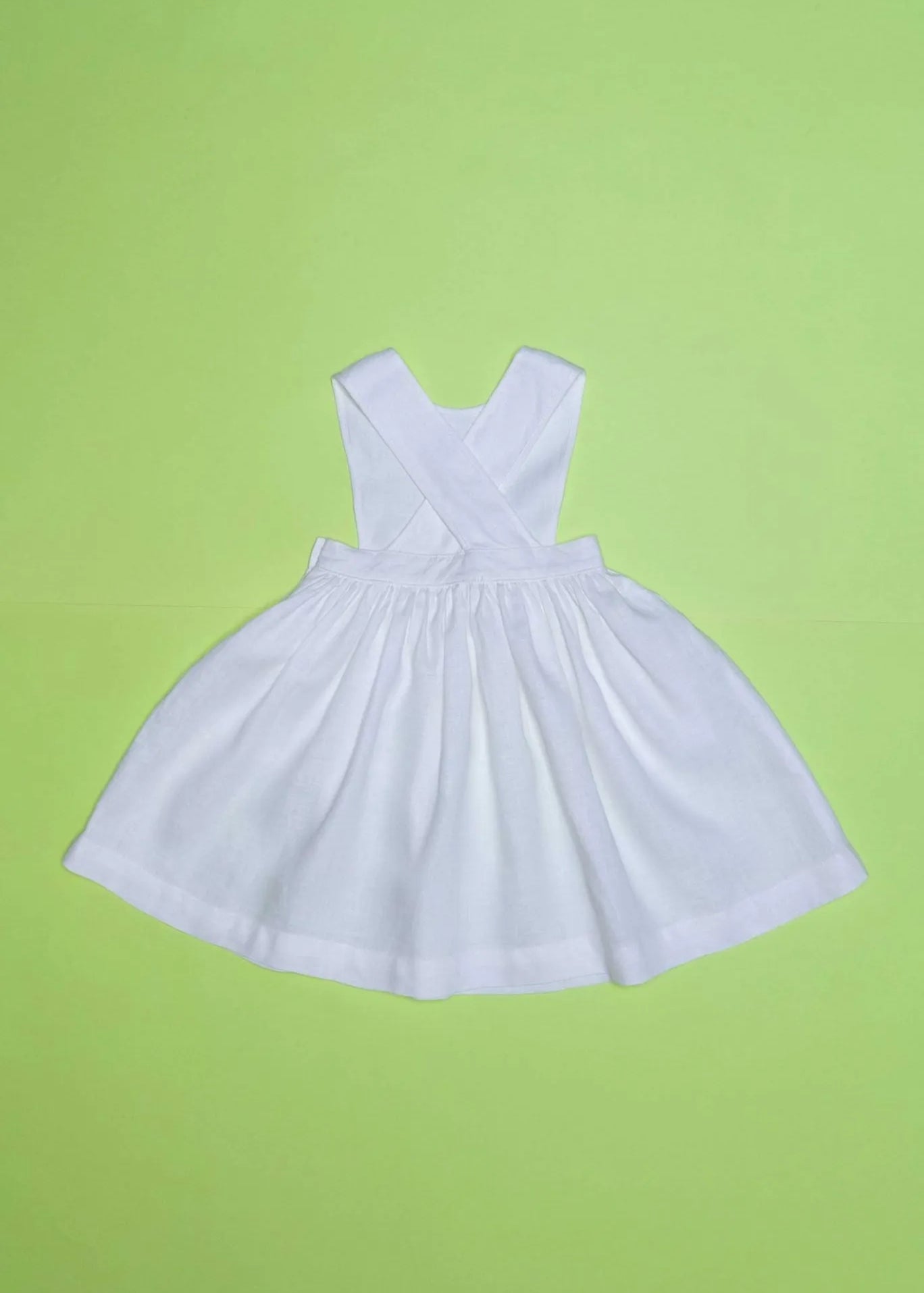 Vista de costas do vestido infantil de linho branco com alças cruzadas e saia ampla, em superfície verde.