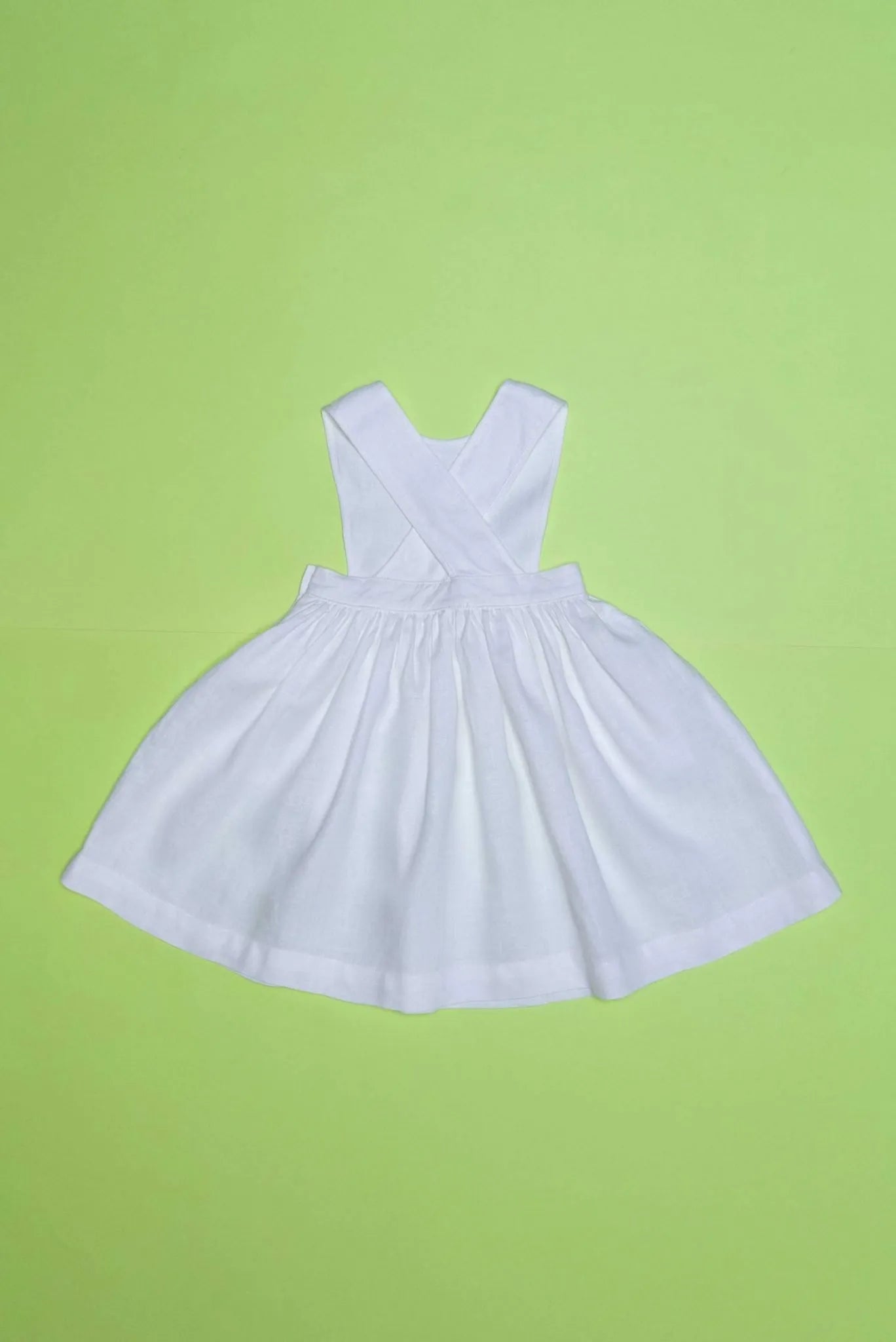 Vista de costas do vestido infantil de linho branco com alças cruzadas e saia ampla, em superfície verde.