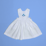 Vestido infantil de linho branco com bordado à mão de conchas azuis para meninas, visto de frente em superfície azul.