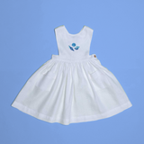 Vestido infantil de linho branco com bordado à mão de conchas azuis para meninas, visto de frente em superfície azul.